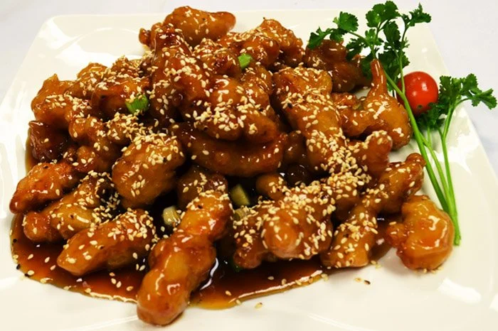 Sweet and sour chicken garnished with sesame seeds, cilantro, cherry tomato, and green onions on a white plate.