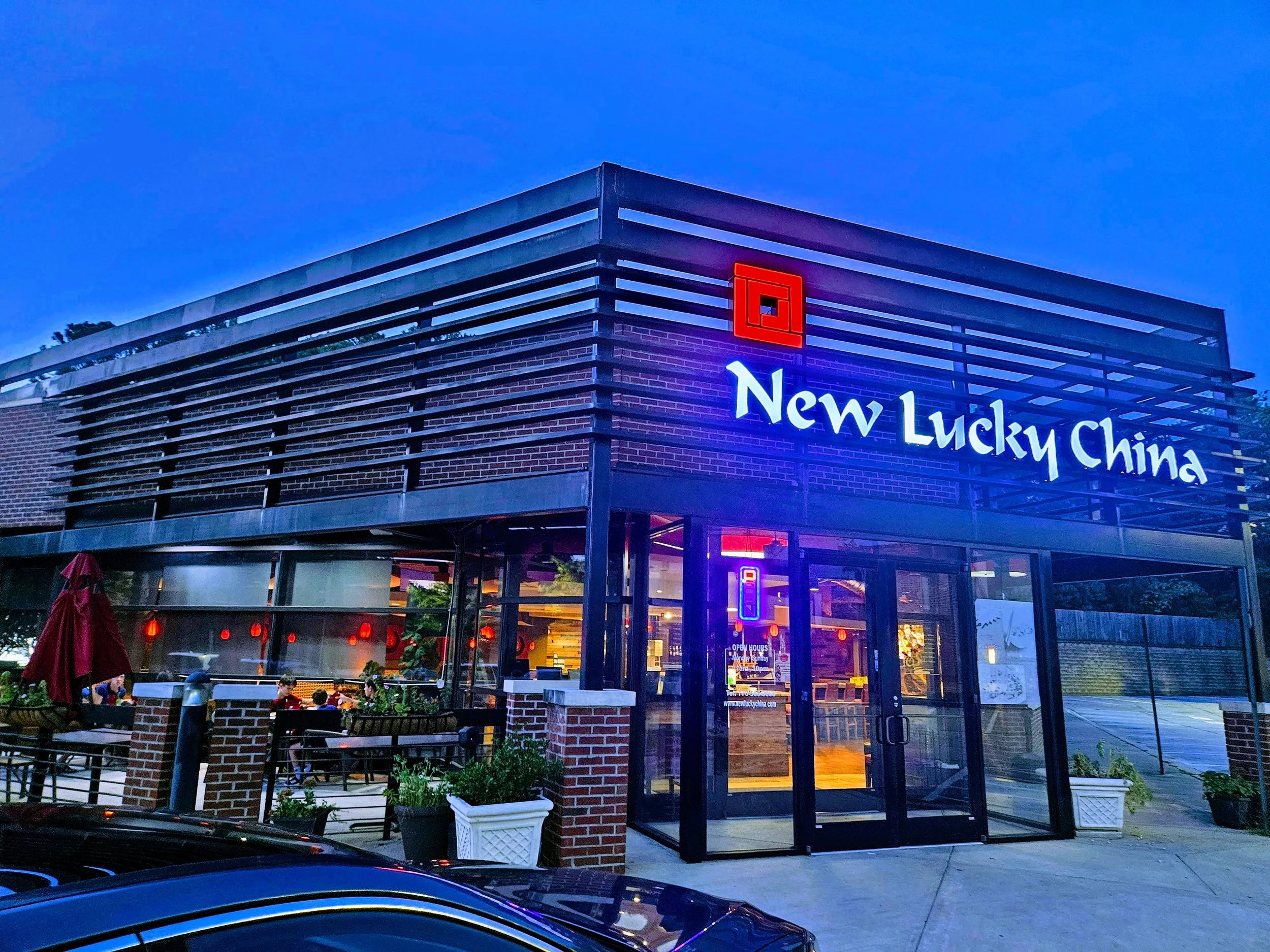 A two-story restaurant building with a modern design, neon signs, outdoor patio seating, and the name 'New Lucky China' illuminated on the front, taken during dusk.