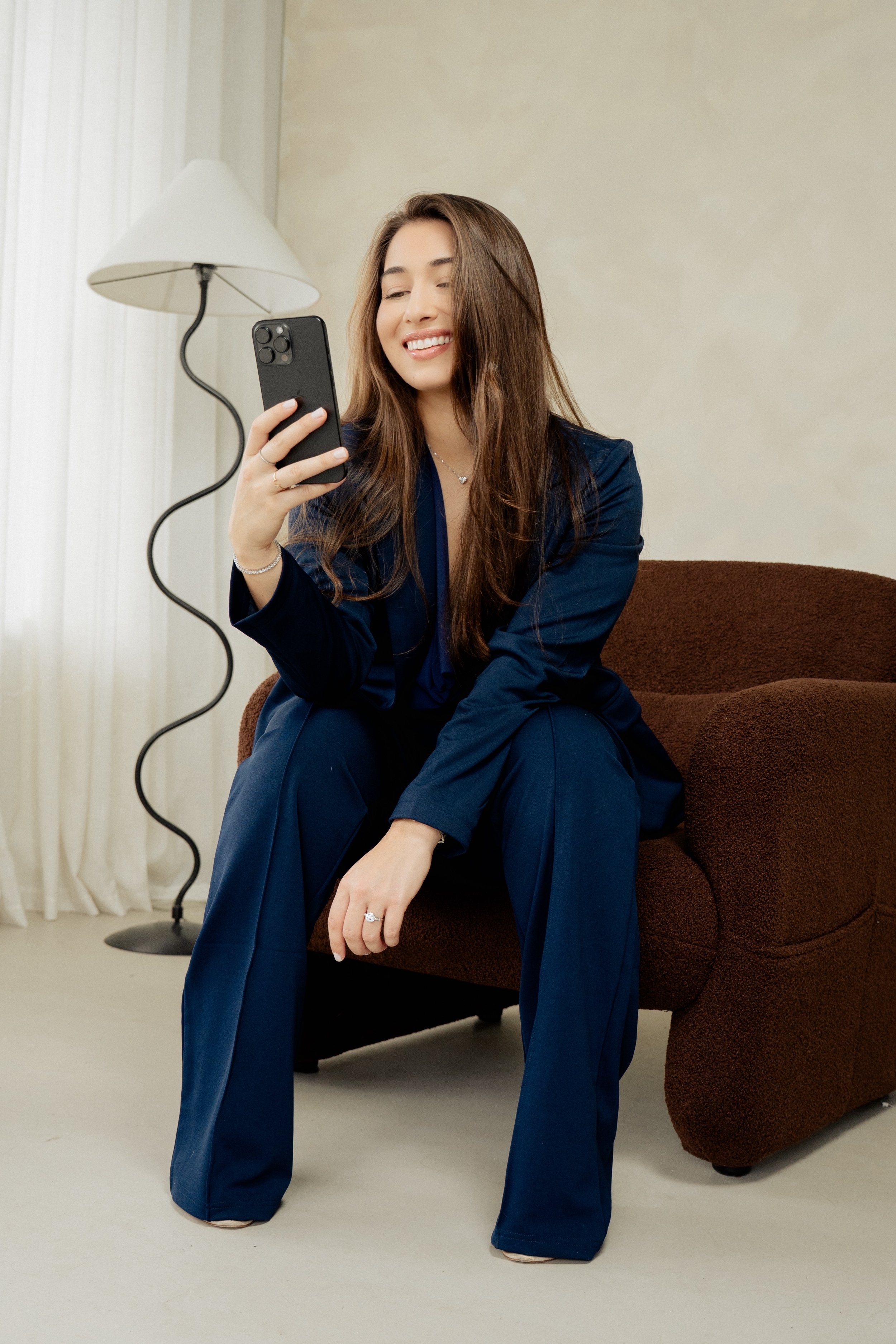 Woman sitting on a brown sofa, smiling while looking at her phone, wearing a navy blue outfit, with a lamp and curtains in the background.