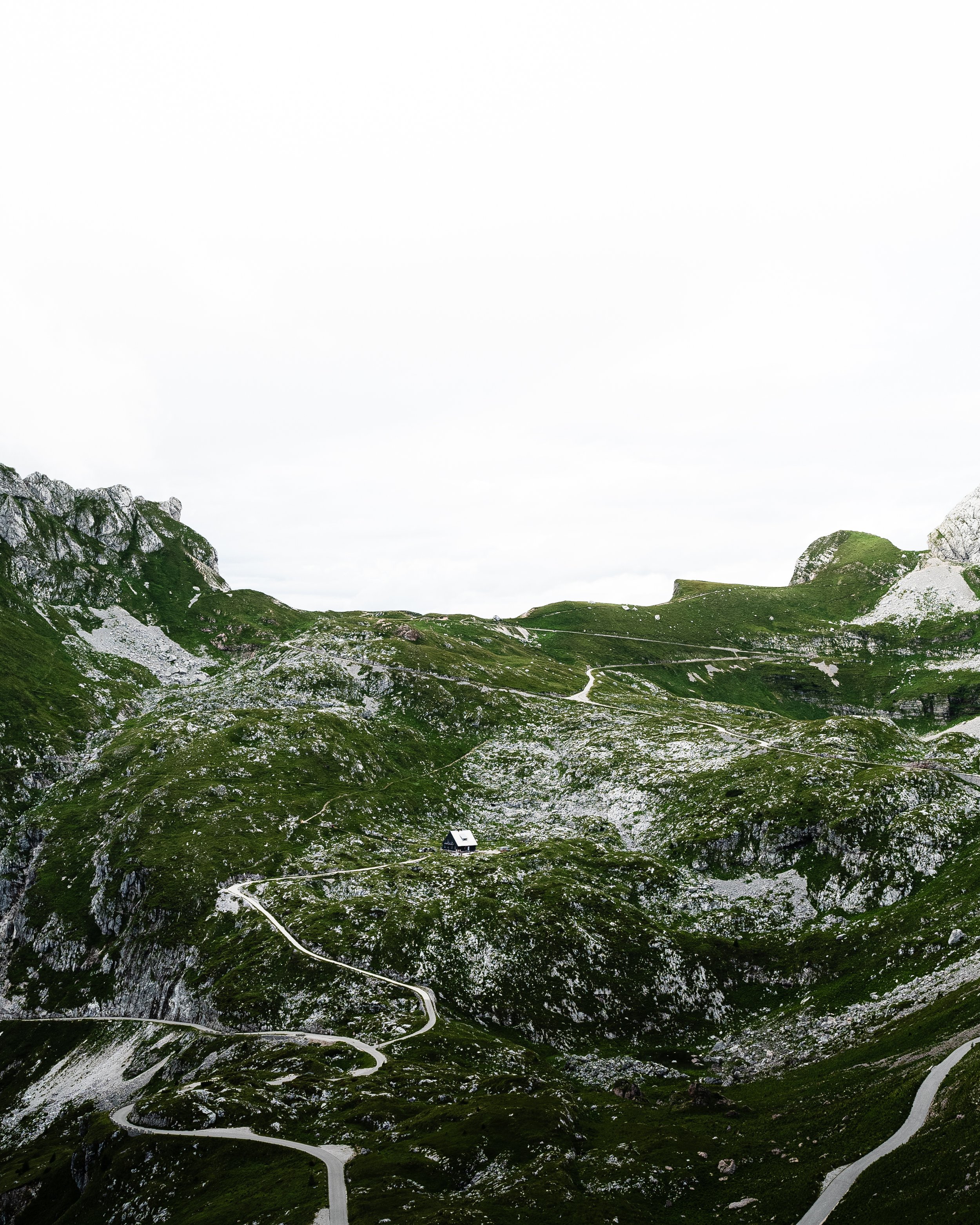 Green mountain landscape with winding paths and a small cabin on a slope under a cloudy sky.