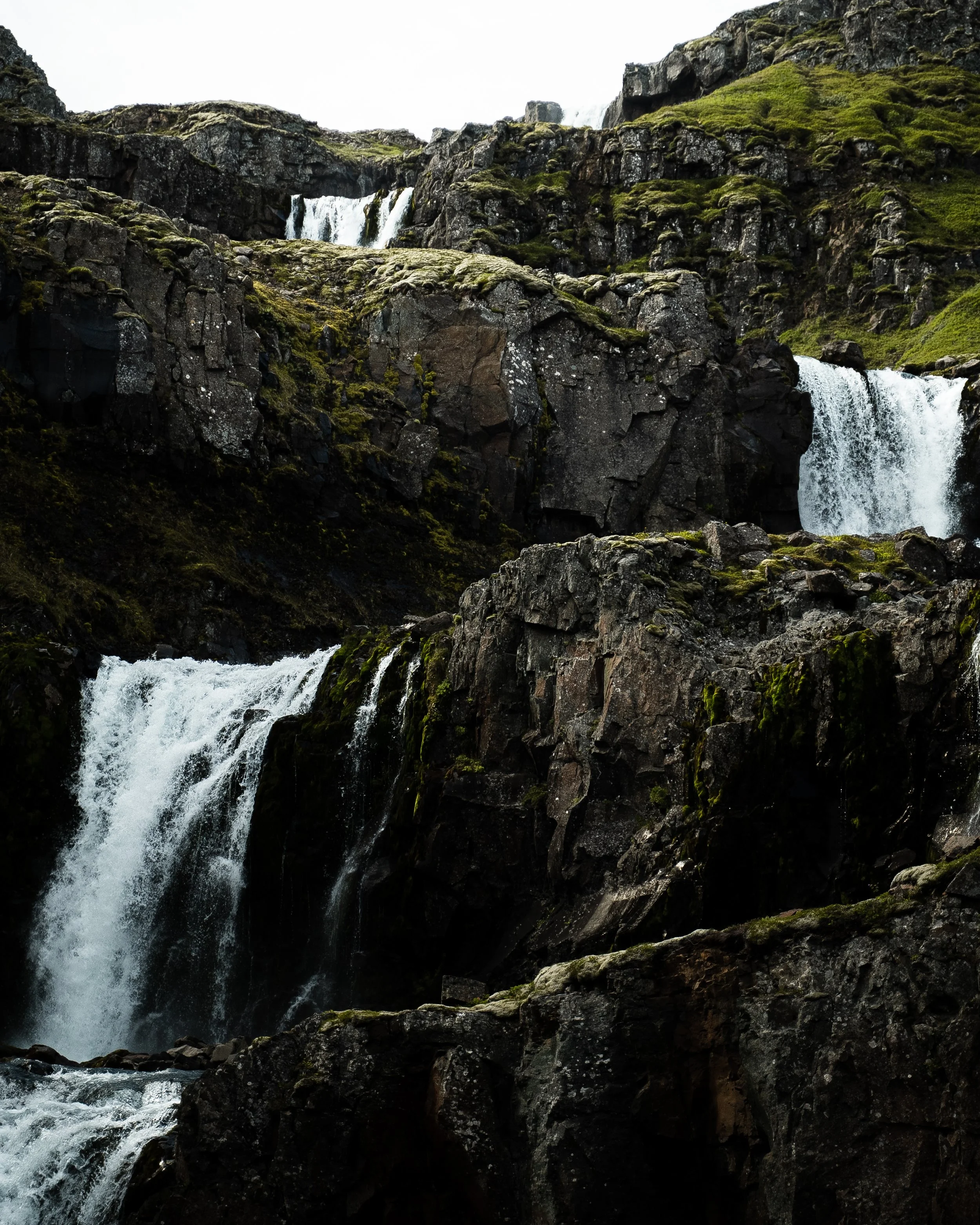 Multiple waterfalls cascading down dark rocky cliffs with patches of green moss in a rugged landscape