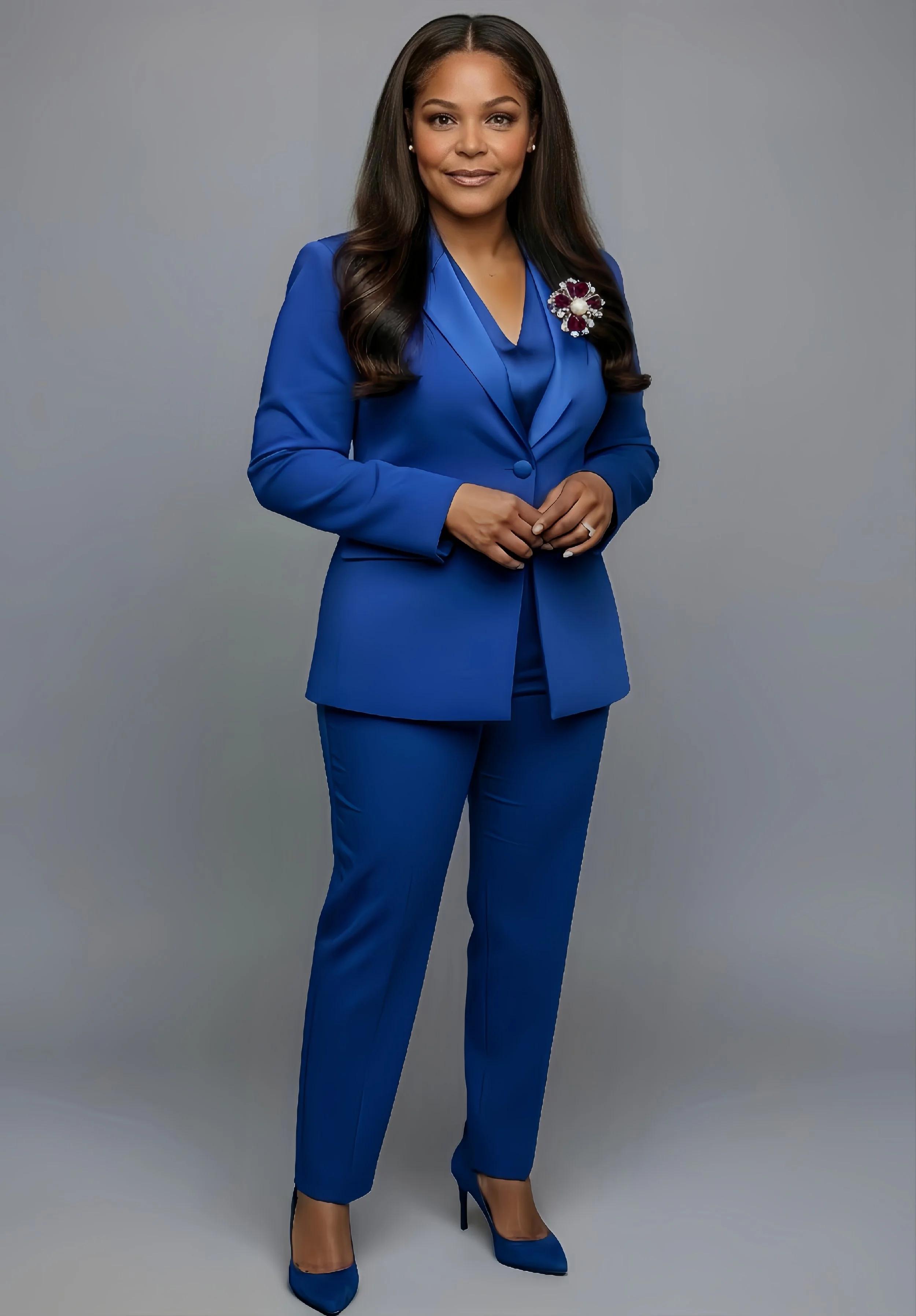 A woman in a blue suit with a decorative brooch, standing against a gray background.
