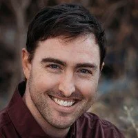 A smiling man with dark hair and a beard, wearing a maroon shirt outside with blurred natural background.
