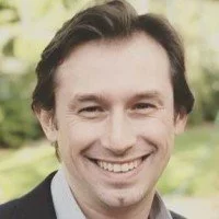 A man smiling outdoors with a blurred green background.