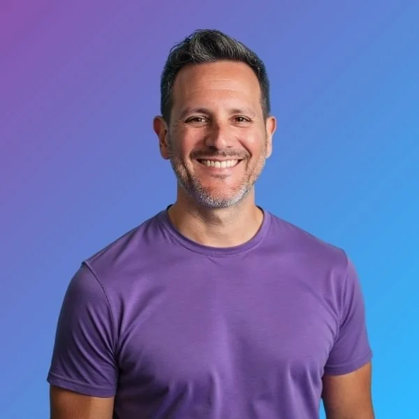 A smiling man with short dark hair and a beard wearing a purple T-shirt against a gradient blue background.