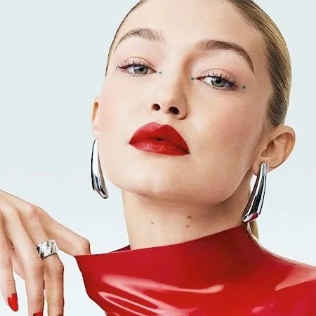 A woman with platinum blonde hair, bold red lipstick, and large silver hoop earrings, wearing a shiny red outfit, looking confident.