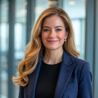 Professional woman with long, wavy red hair wearing a navy blazer and black top, standing in an office environment with glass windows.