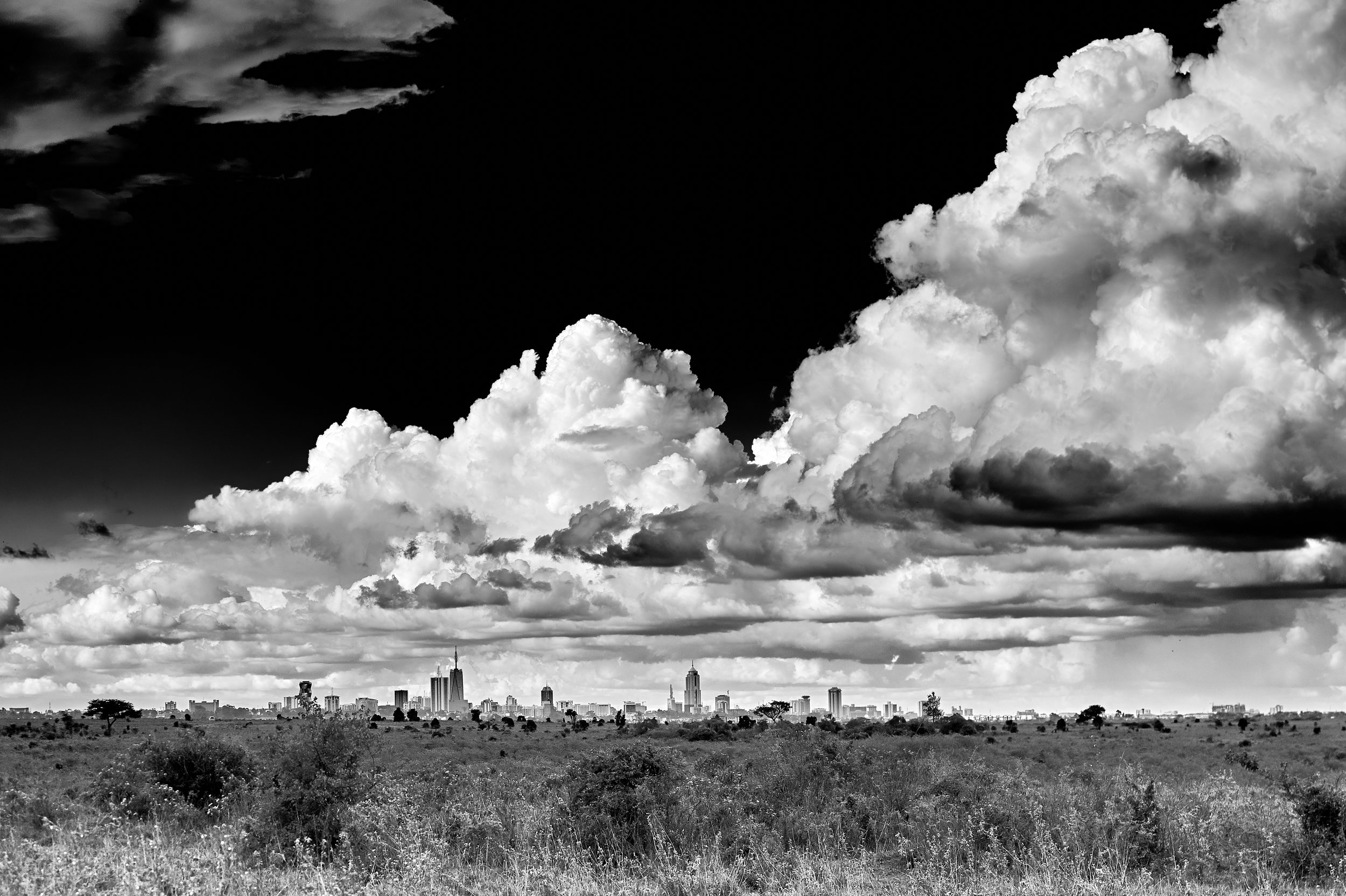 Black and white photo of a city skyline in the distance under a sky filled with large, dramatic clouds.