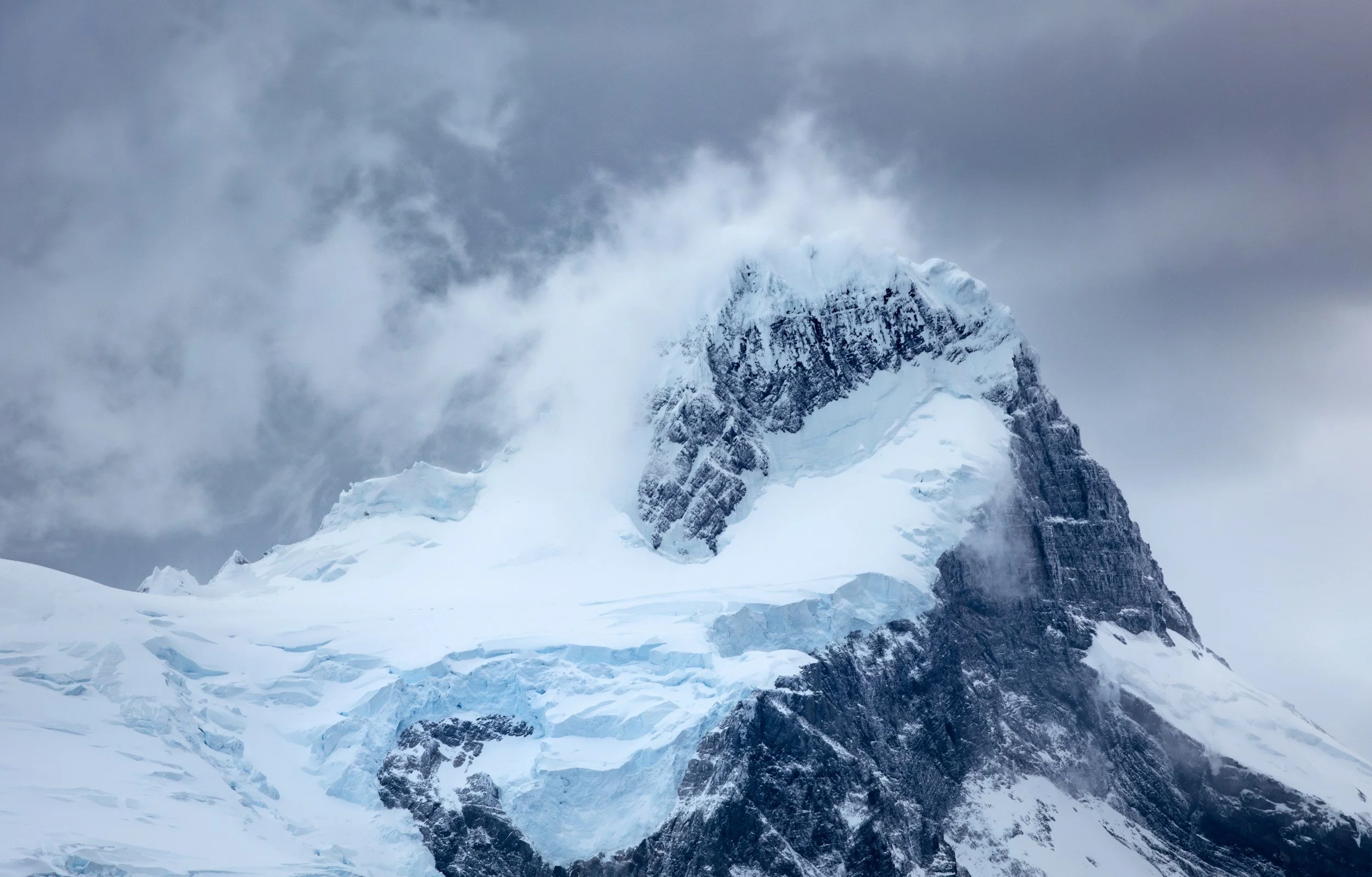 Snow-covered mountain peak with rocky cliffside shrouded in clouds and mist.