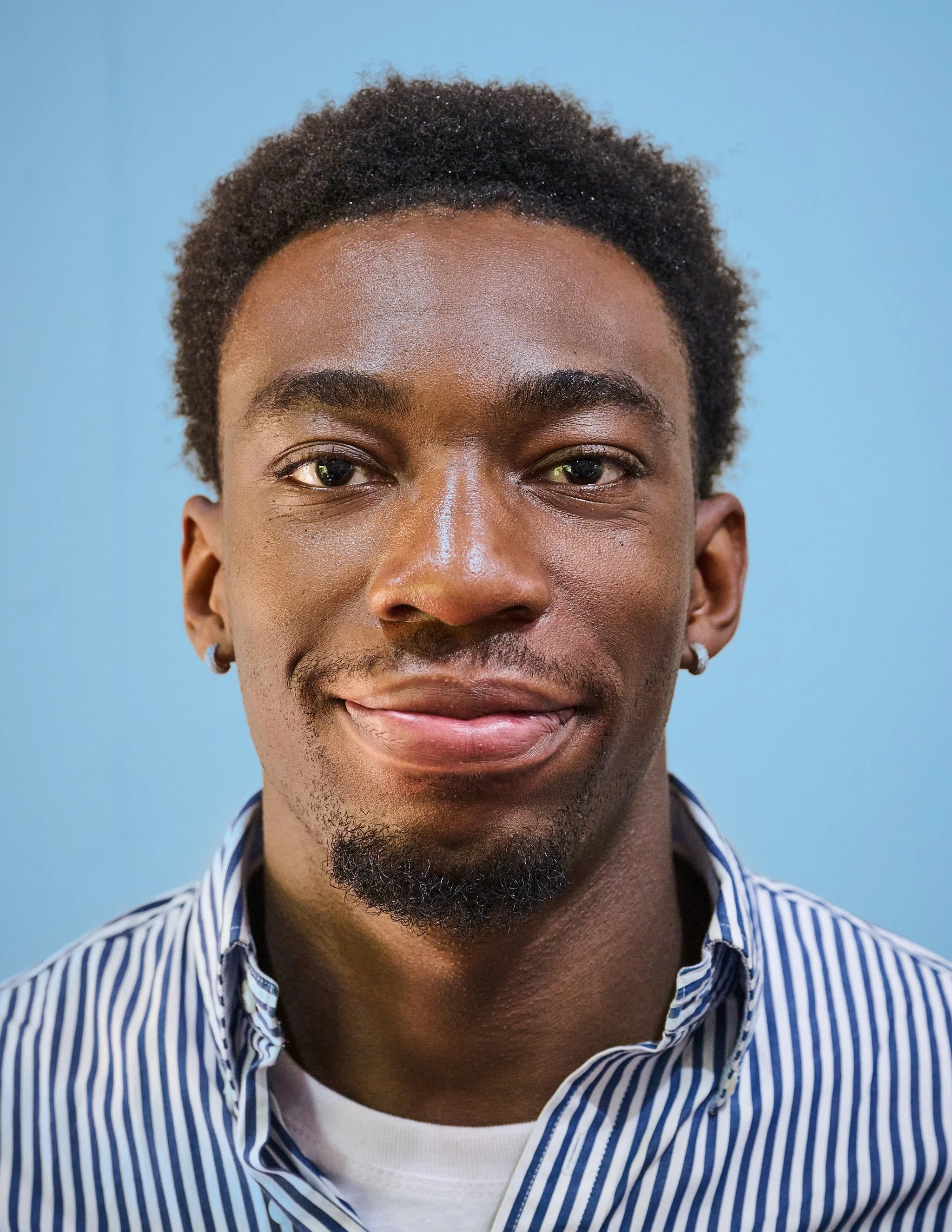 Close-up portrait of a young Black man with short curly hair, earrings, slight beard, wearing a striped shirt and white t-shirt, smiling in front of a blue background.