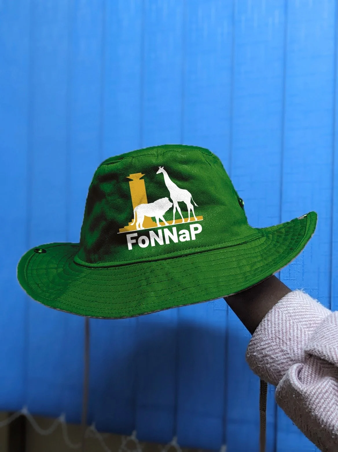 Green hat with the logo of FoNNap, featuring a giraffe and a rhinoceros, being held by a person against a blue background.
