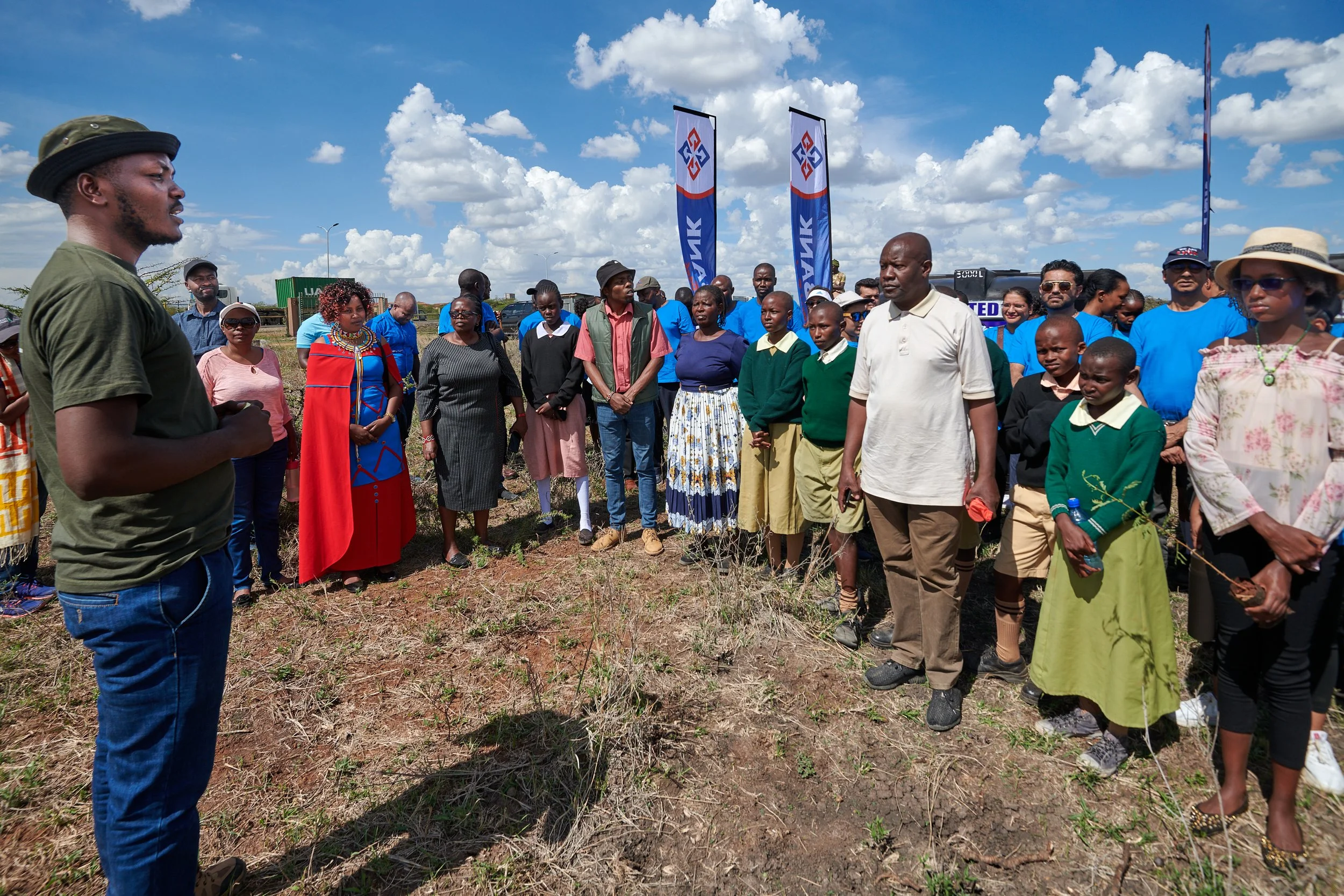 A man in a green shirt and hat speaking to a group of people outdoors on a sunny day, with flags and a truck in the background.