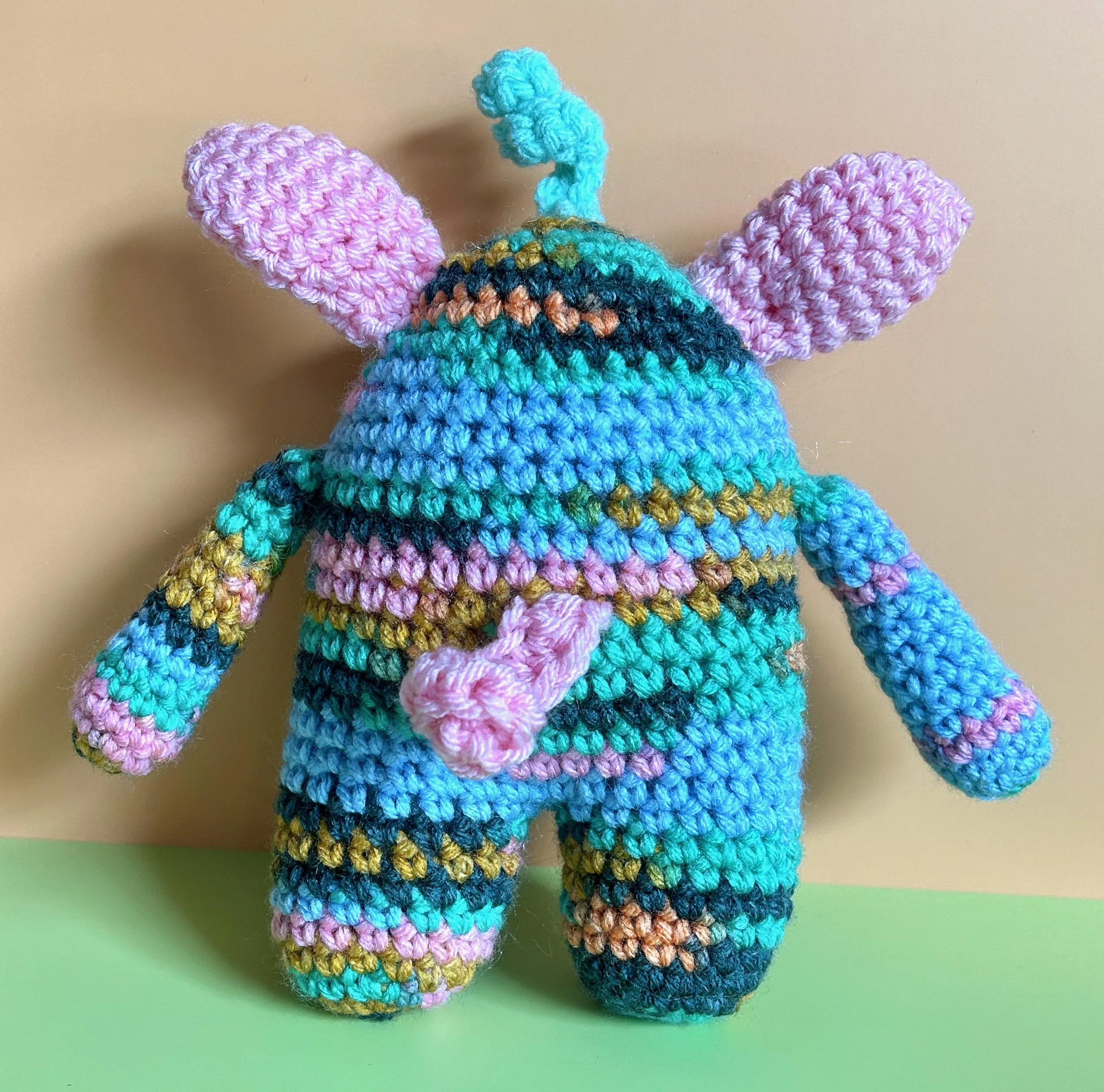 Springy back of light blue, pink, green, and gold monster with pink bunny ears squiggly hair and curly pink tail