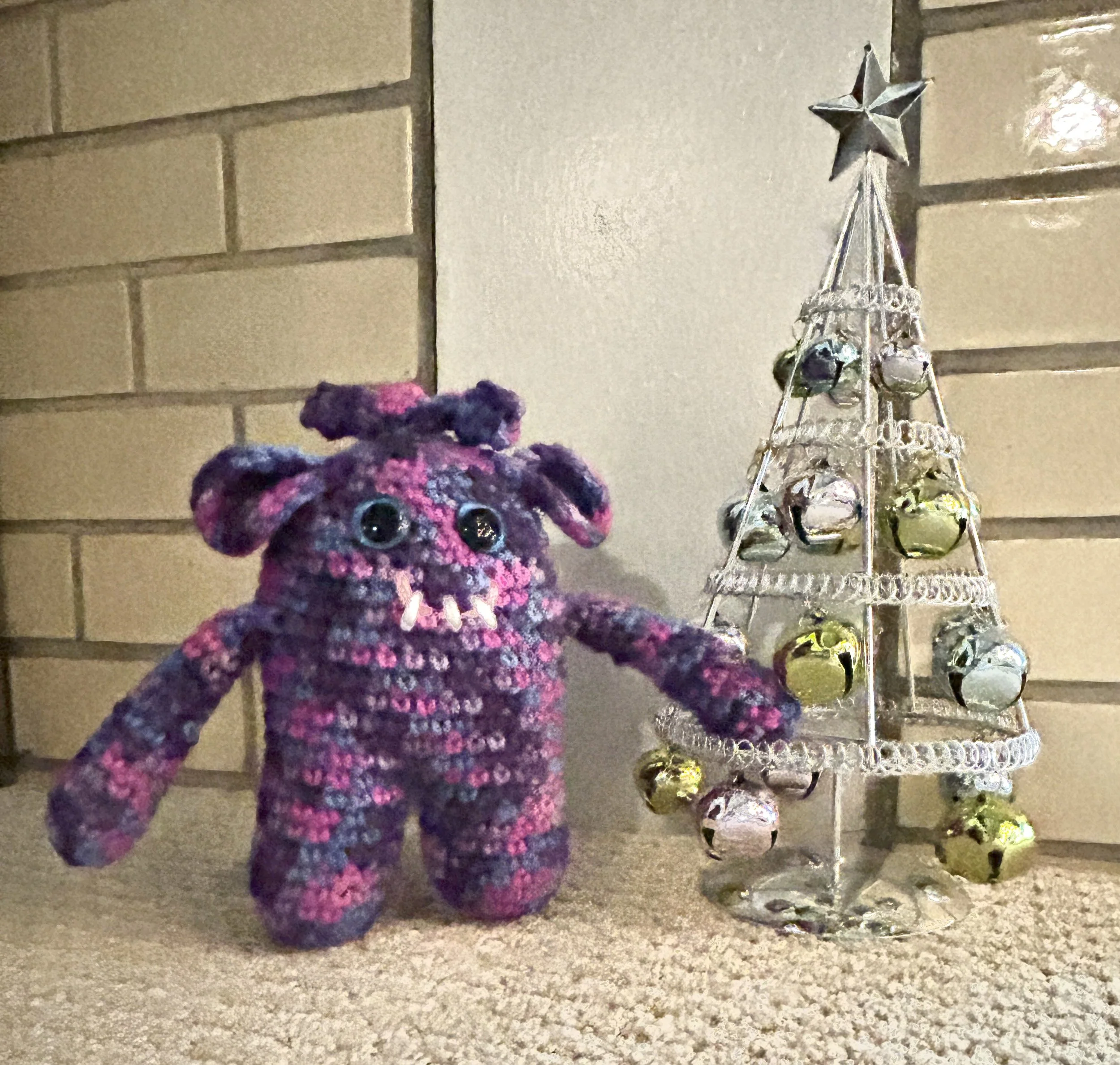 Beryl shades of purple and bright pink monster with blue googly eyes smiling with 3 teeth trimming a silver Christmas tree with pastel colored bells and a star