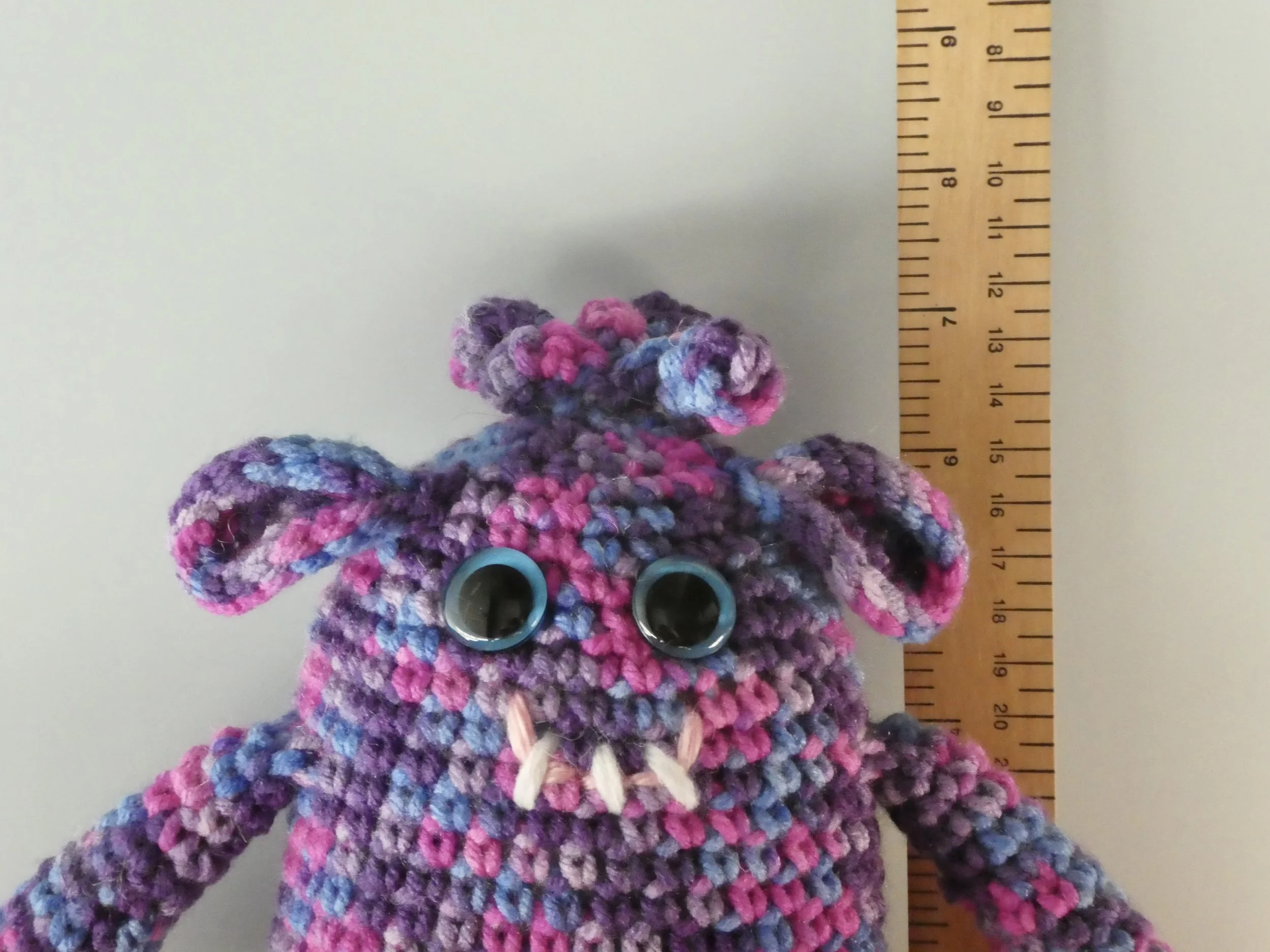 Beryl closeup of face shades of purple and bright pink monster with blue googly eyes smiling with 3 teeth standing next to ruler body about 6-1/2 inches tall