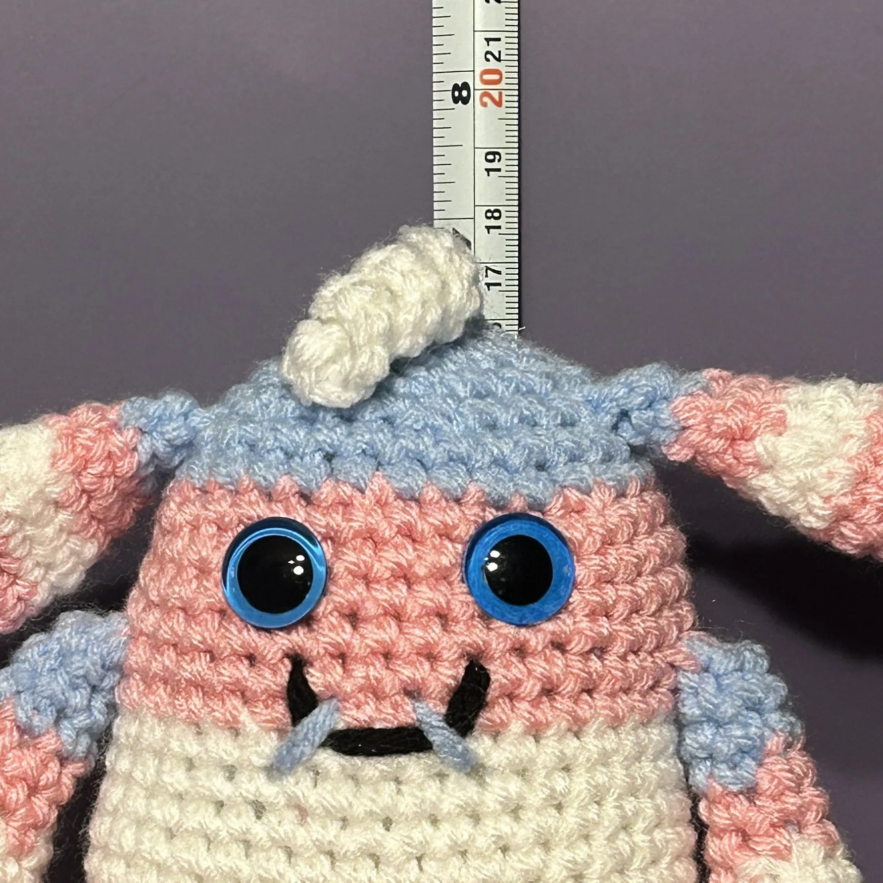 Nicci light blue pink and white striped monster with bunny ears and one white white squiggle of hair, blue googly eyes, smiling with 2 teeth in front of ruler, body about 6-3/4 inches tall