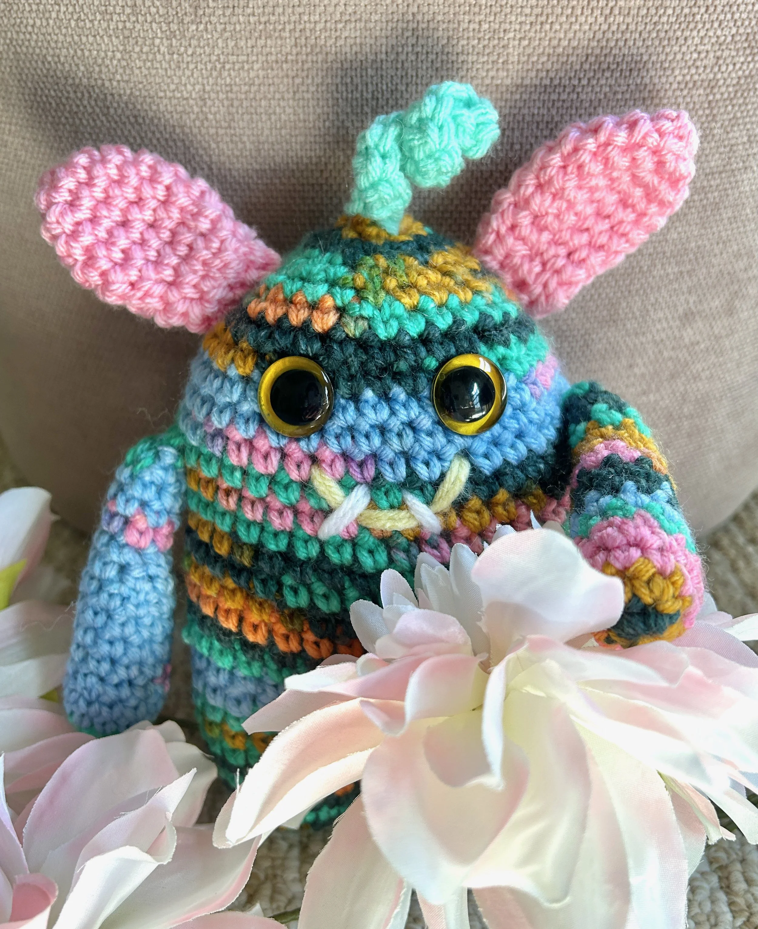 Springy light blue, pink, green, and gold monster with pink bunny ears squiggly hair gold googly eyes smiling with 2 teeth nestled in large light pink flowers