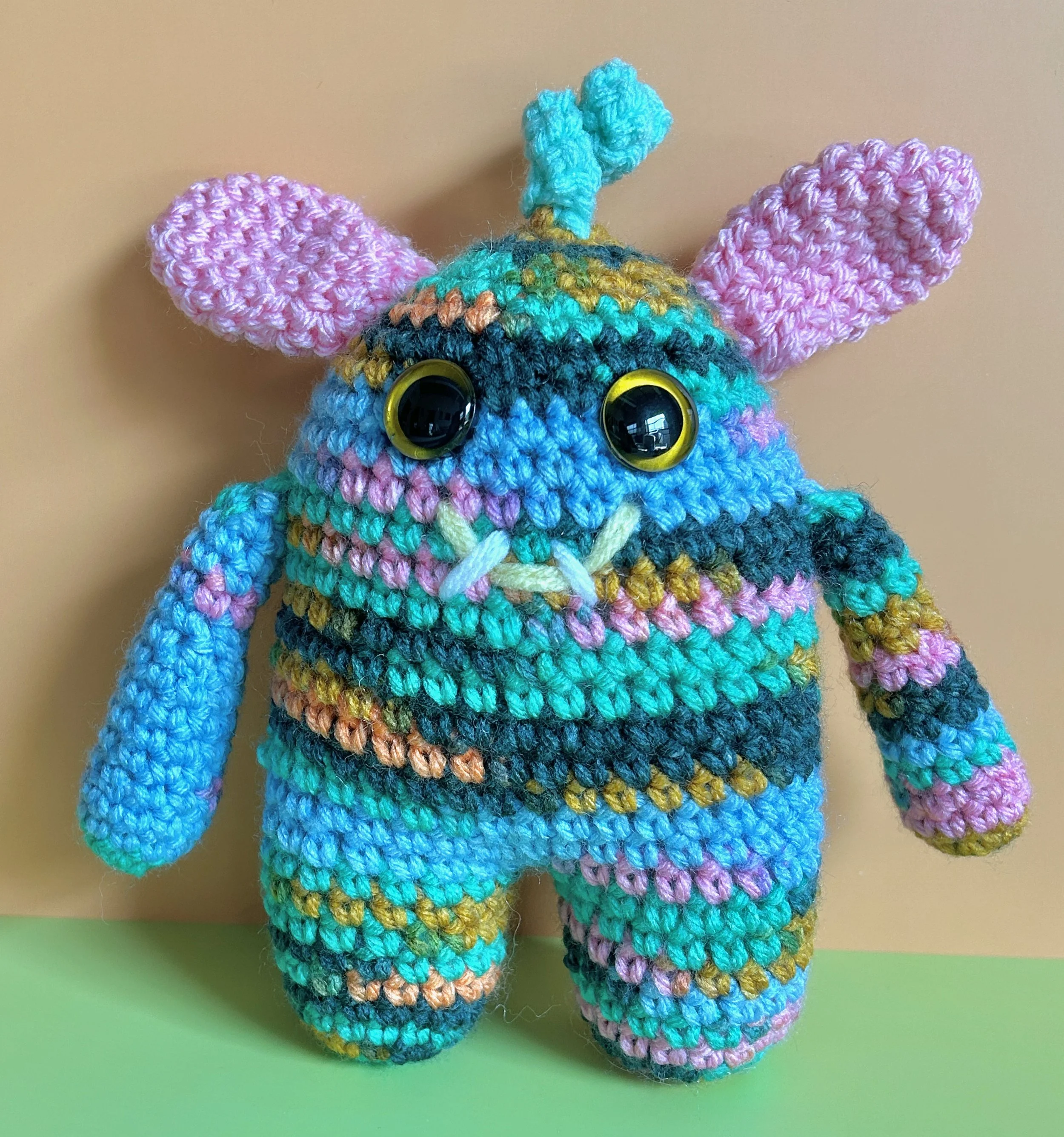 Springy light blue, pink, green, and gold monster with green squiggly hair pink bunny ears gold googly eyes smiling with 2 teeth