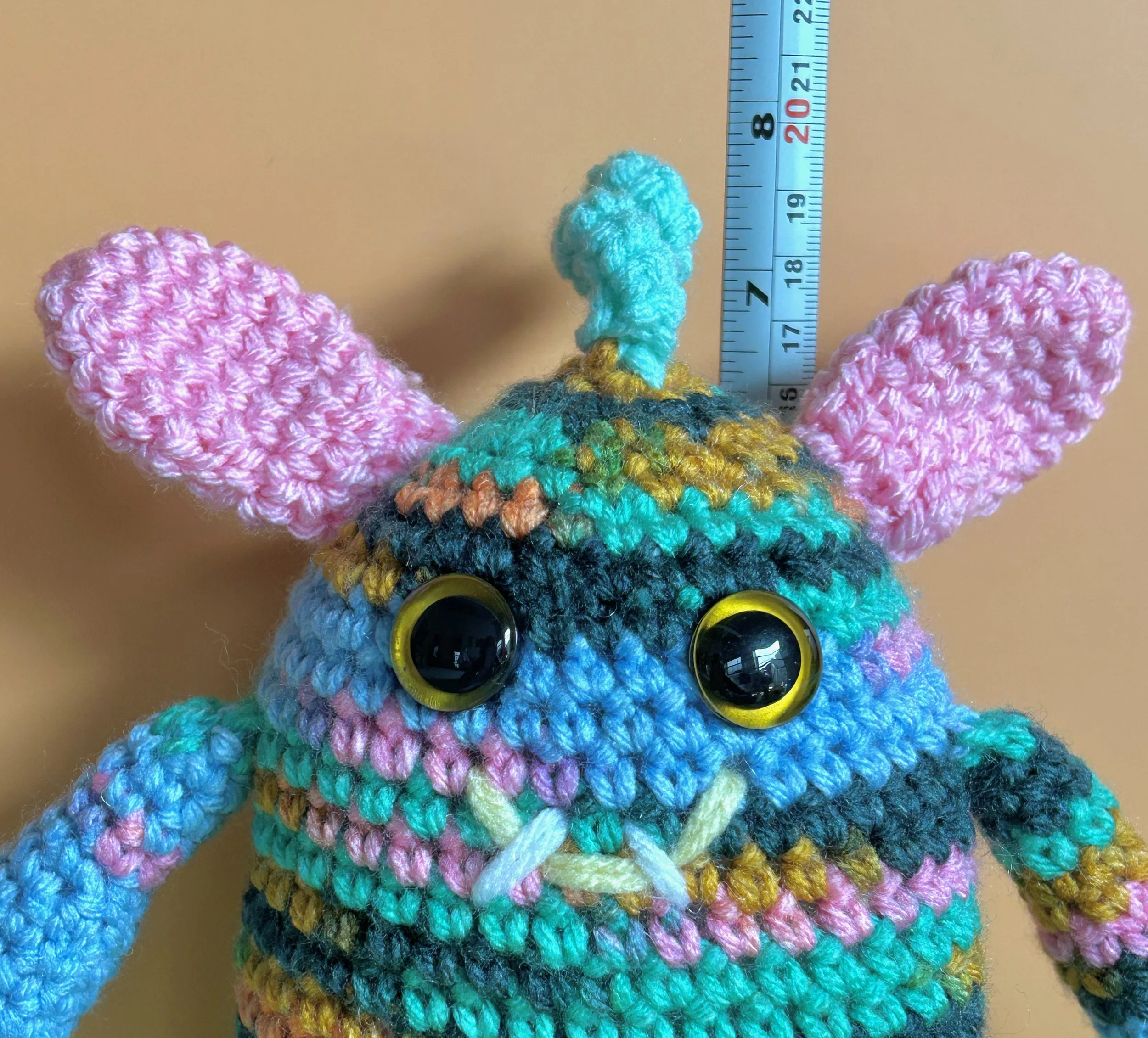 Springy closeup of face light blue, pink, green, and gold monster with pink bunny ears squiggly hair gold googly eyes smiling with 2 teeth standing in front of ruler body about 6-1/2 inches tall