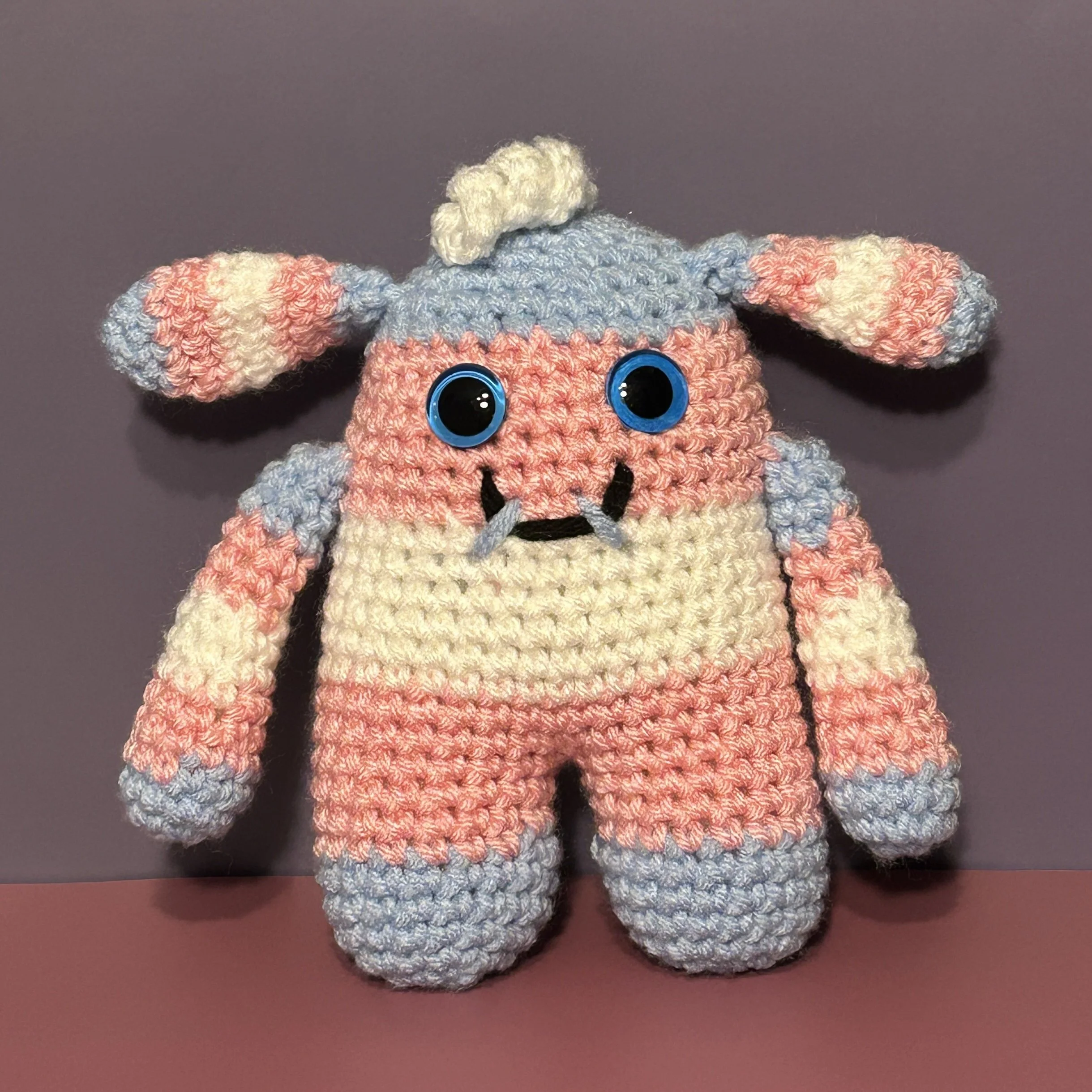 Nicci light blue pink and white striped monster with bunny ears and a one white squiggle of hair, blue googly eyes, smiling with 2 teeth