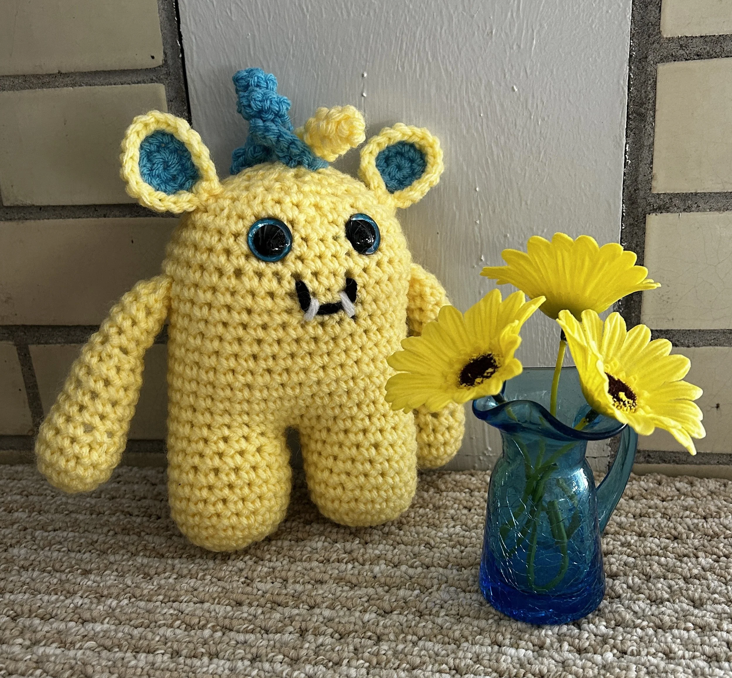 Flora daffodil yellow monster with blue squiggly hair, blue googly eyes, smiling with 2 teeth admiring a blue glass pitcher style vase with three yellow brown eyed daisies