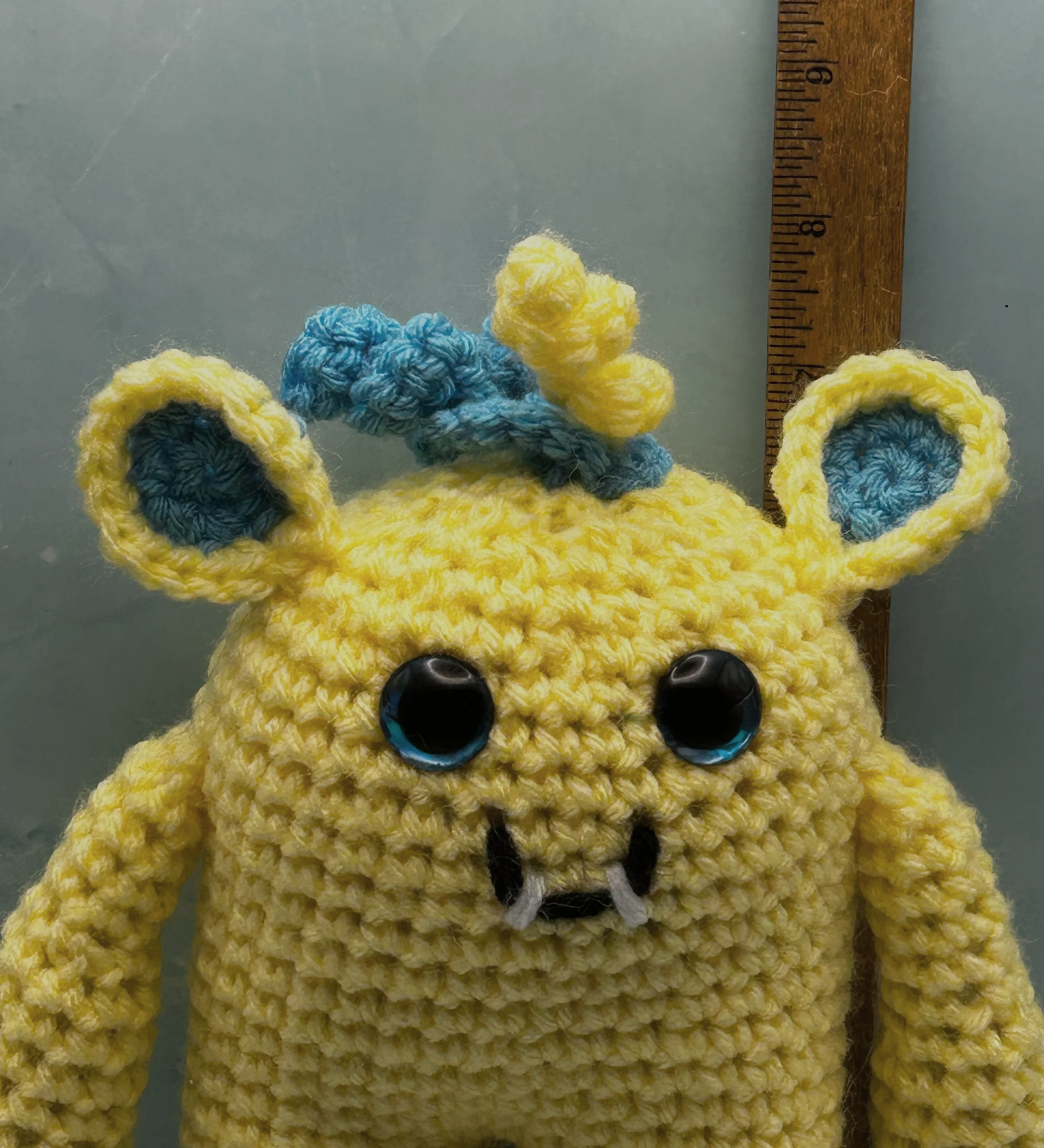Flora closeup of face daffodil yellow monster, blue squiggly hair, blue googly eyes, smiling with 2 teeth next to ruler body about 6-1/2 inches tall