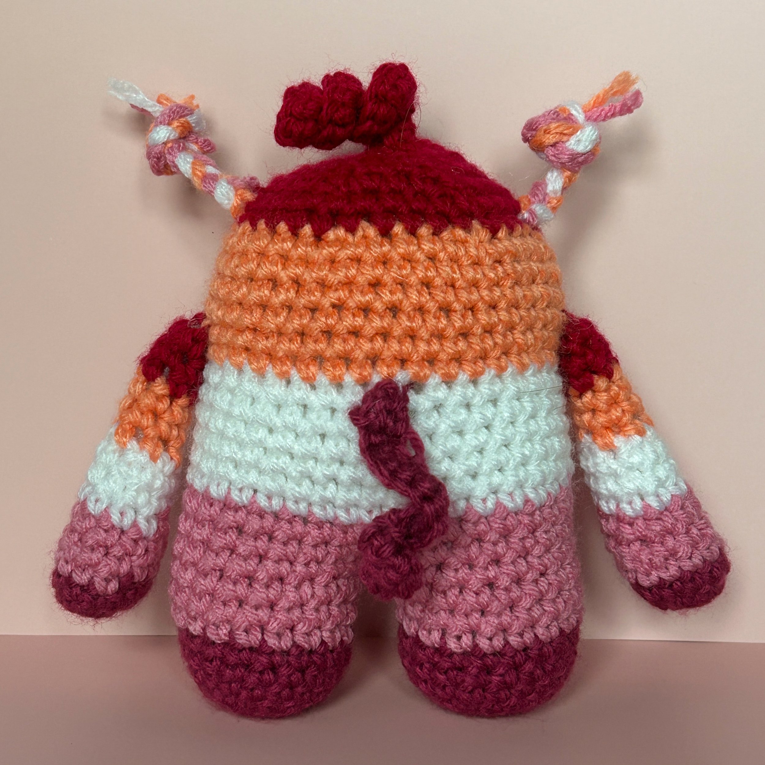 back of dark orange, light orange, white, light pink, dark pink striped monster with braids and a squiggle hair and curly dark pink tail