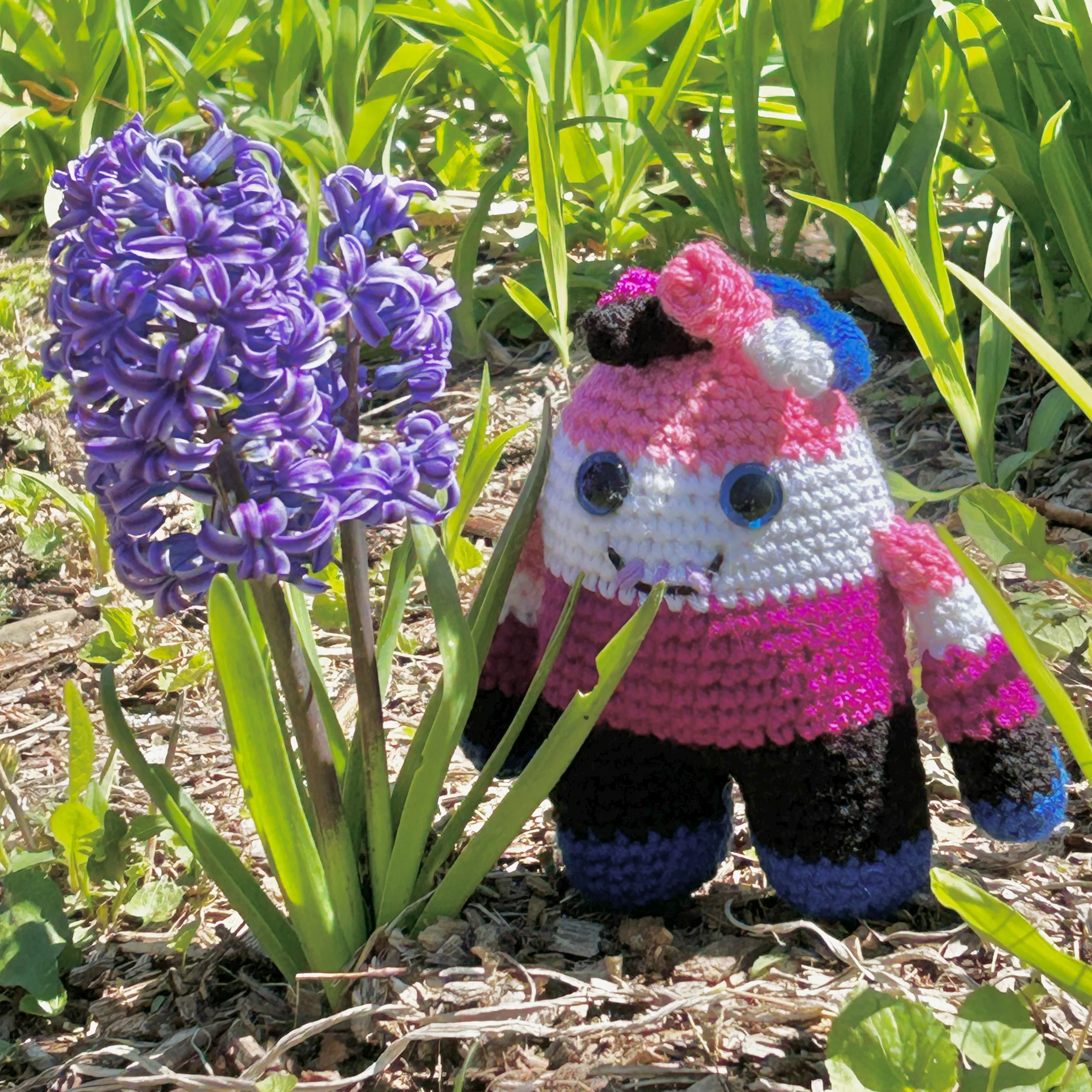 friendly pink, white, purple, black, and blue striped monster with squiggly hair blue googly eyes smiling with 3 teeth next to a purple hyacinth and surrounded by green plant stems