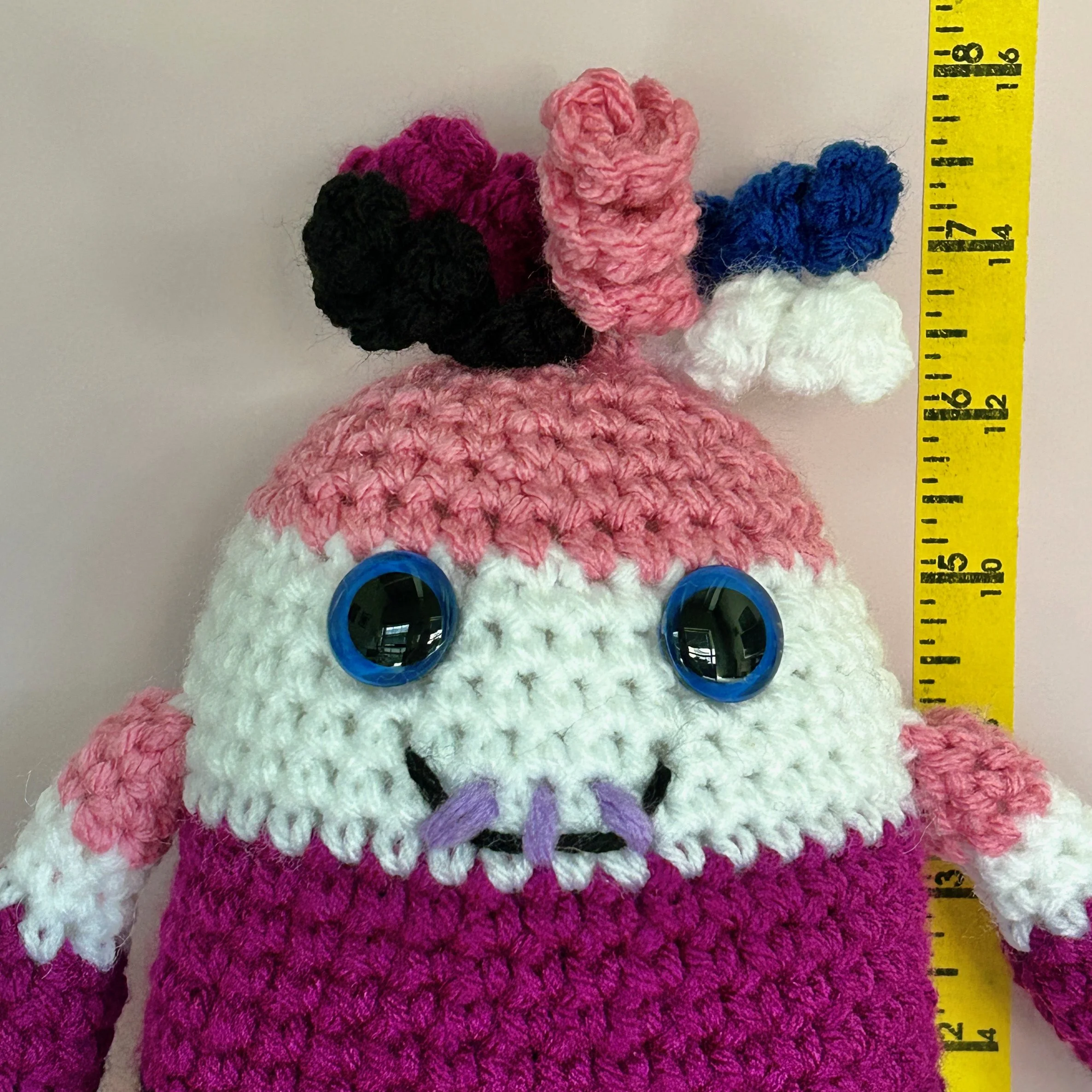 closeup of friendly pink, white, purple, black, and blue striped monster with squiggly hair blue googly eyes smiling with 3 teeth standing in front of ruler body about 6-1/2 inches tall