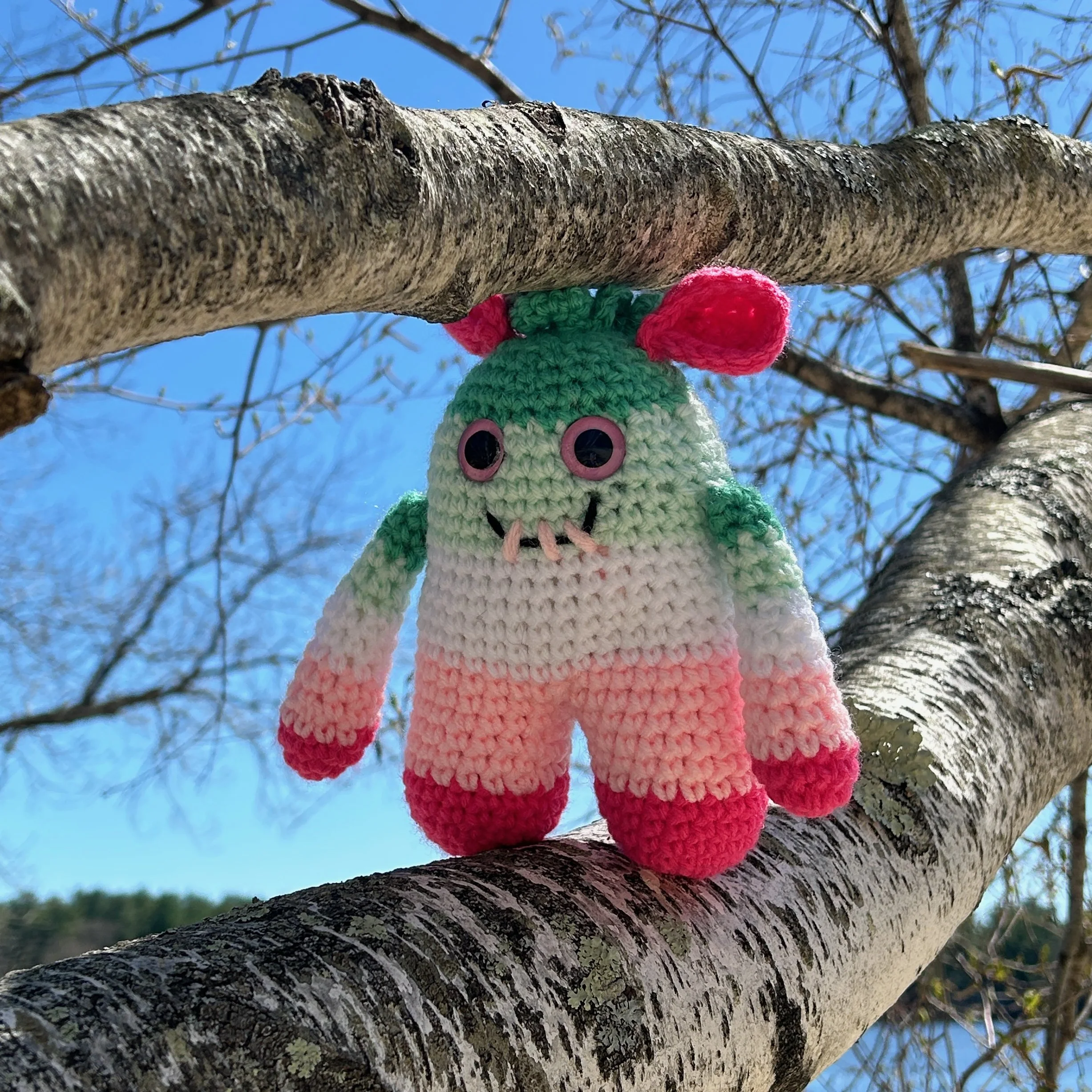 friendly green, white, and pink striped monster with bright pink folded ears squiggly jade green hair standing between 2 limbs of a tree with a bright blue sky in the background