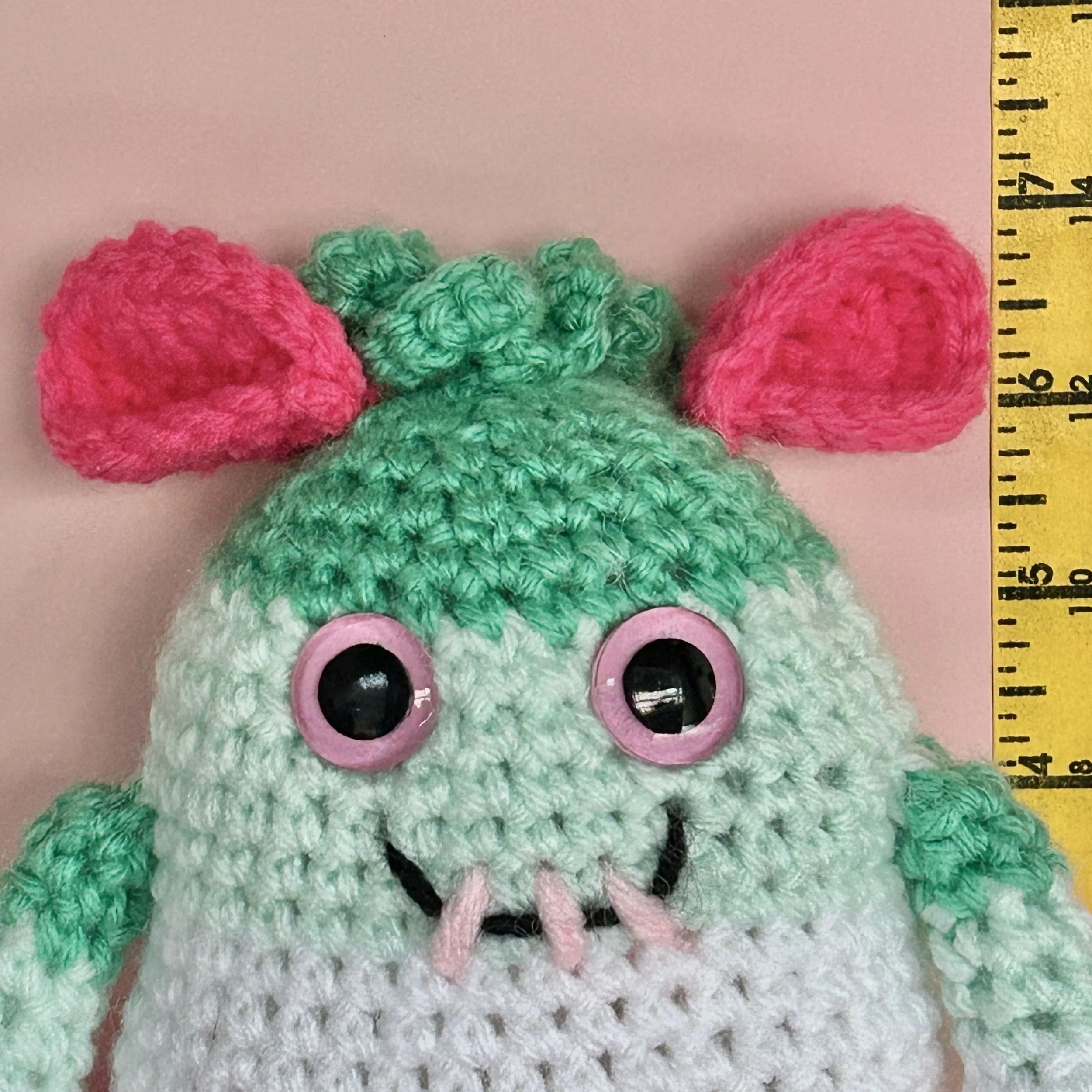 closeup of friendly green, white, and pink striped monster with bright pink folded ears squiggly jade green hair pink googly eyes smiling with 3 teeth standing next to ruler body about 6 inches tall