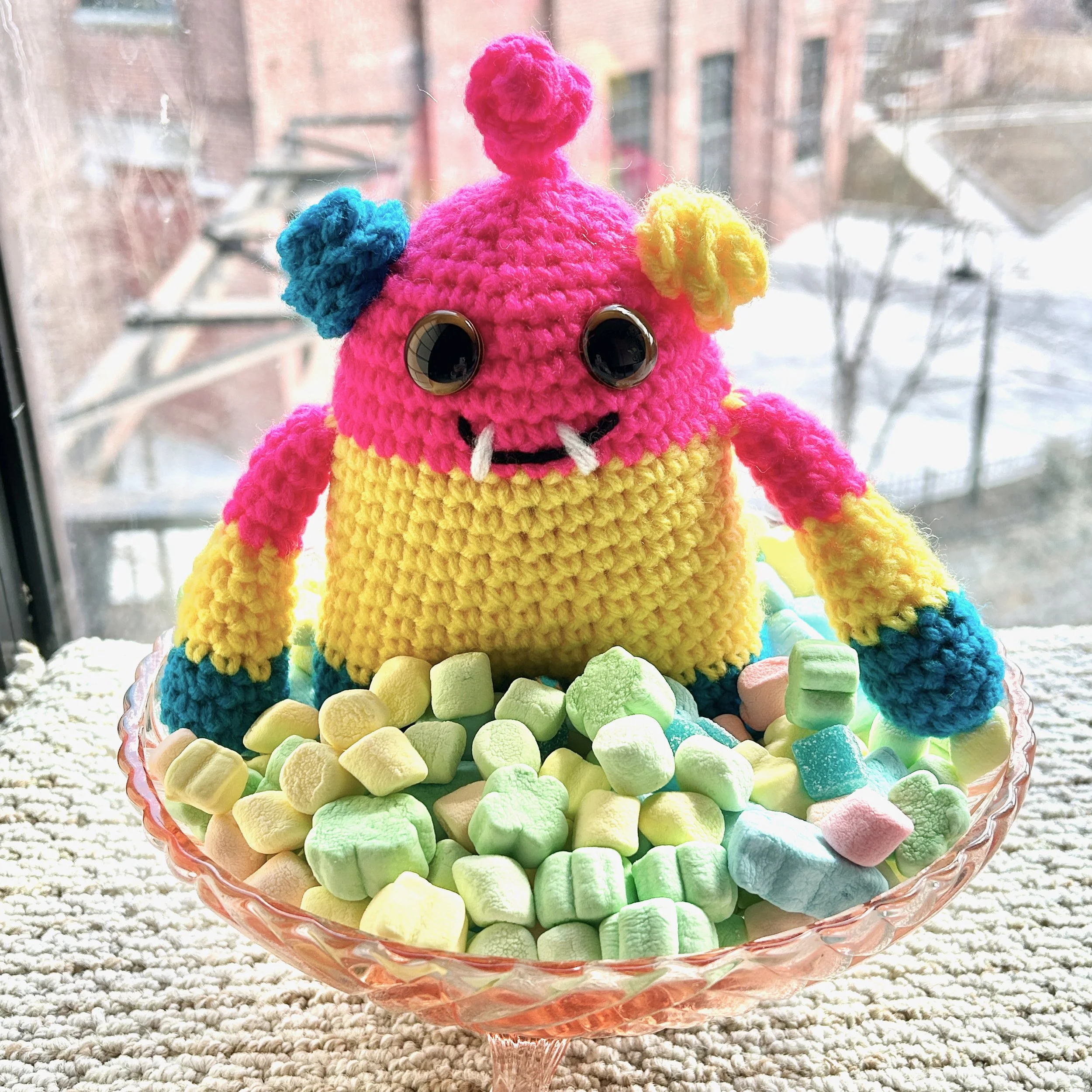 friendly bright pink yellow and blue striped monster with curly ears and pink squiggle hair yellow googly eyes smiling with two teeth in a pink glass bowl filled with pastel marshmallows