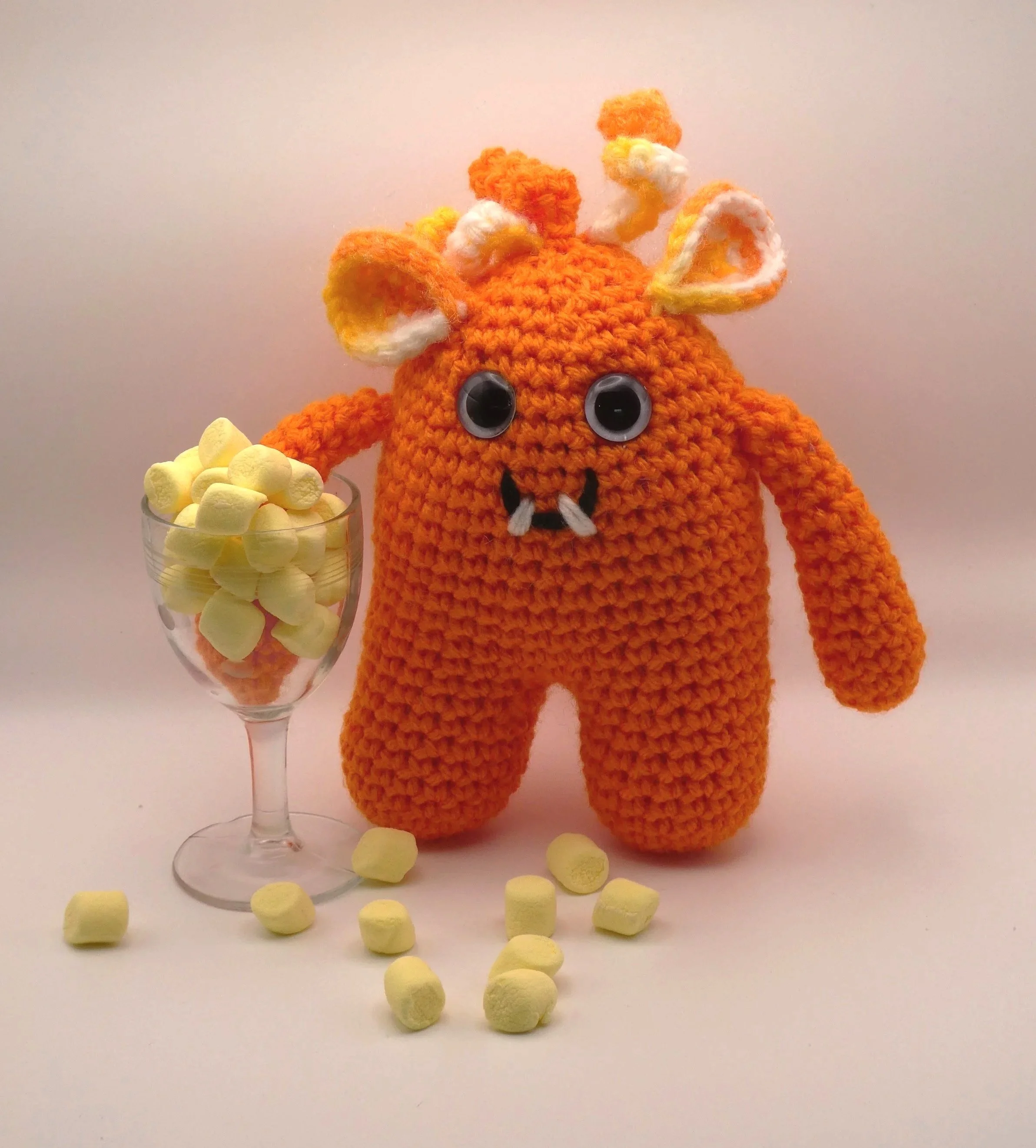 A crocheted orange monster with large googly eyes, horns, and a mouth with two white teeth, holding a glass filled with yellow marshmallows, with marshmallows scattered on the white surface around it.