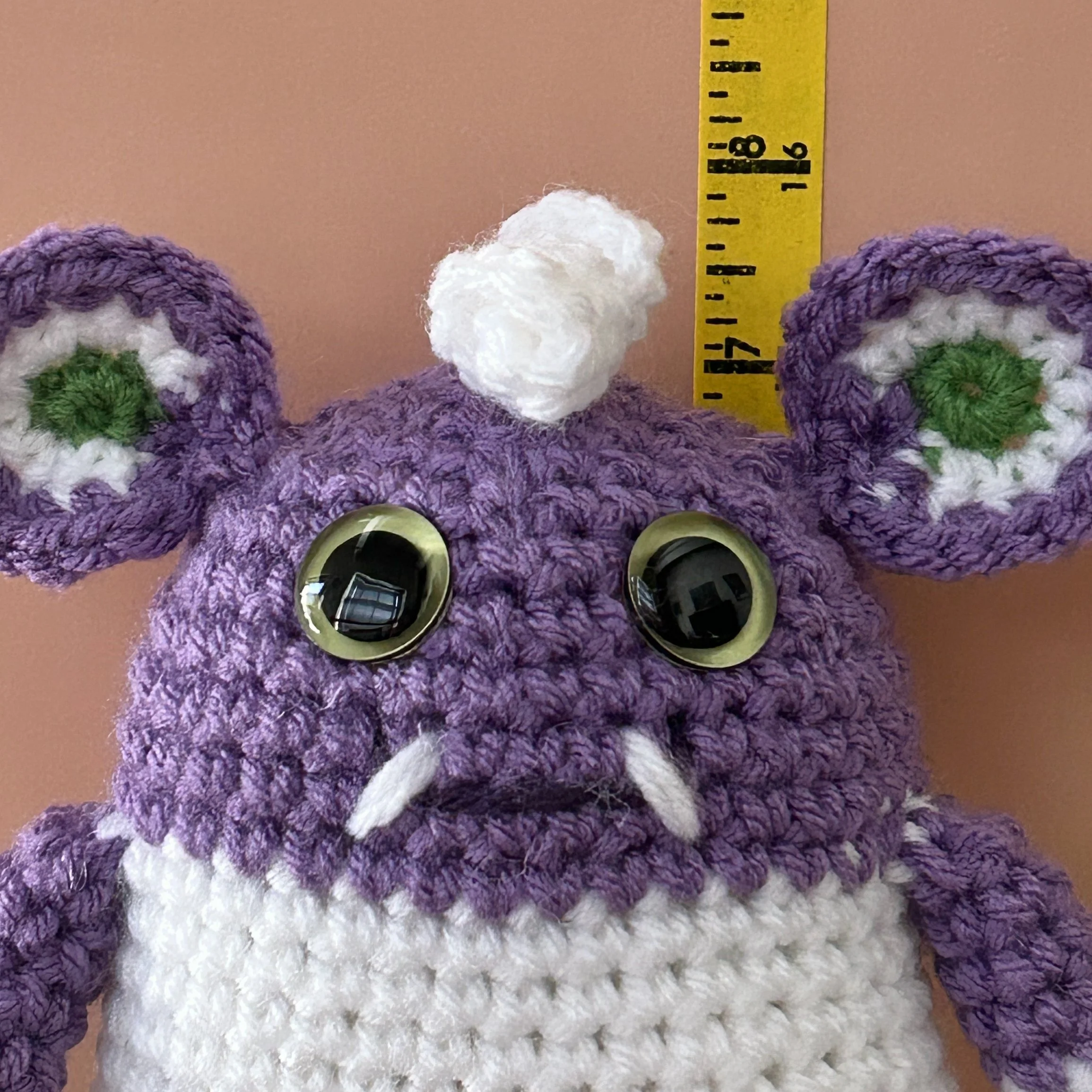 closeup of friendly purple white and green striped monster with round ears light green googly eyes smiling with 2 teeth next to ruler body about 6 inches tall