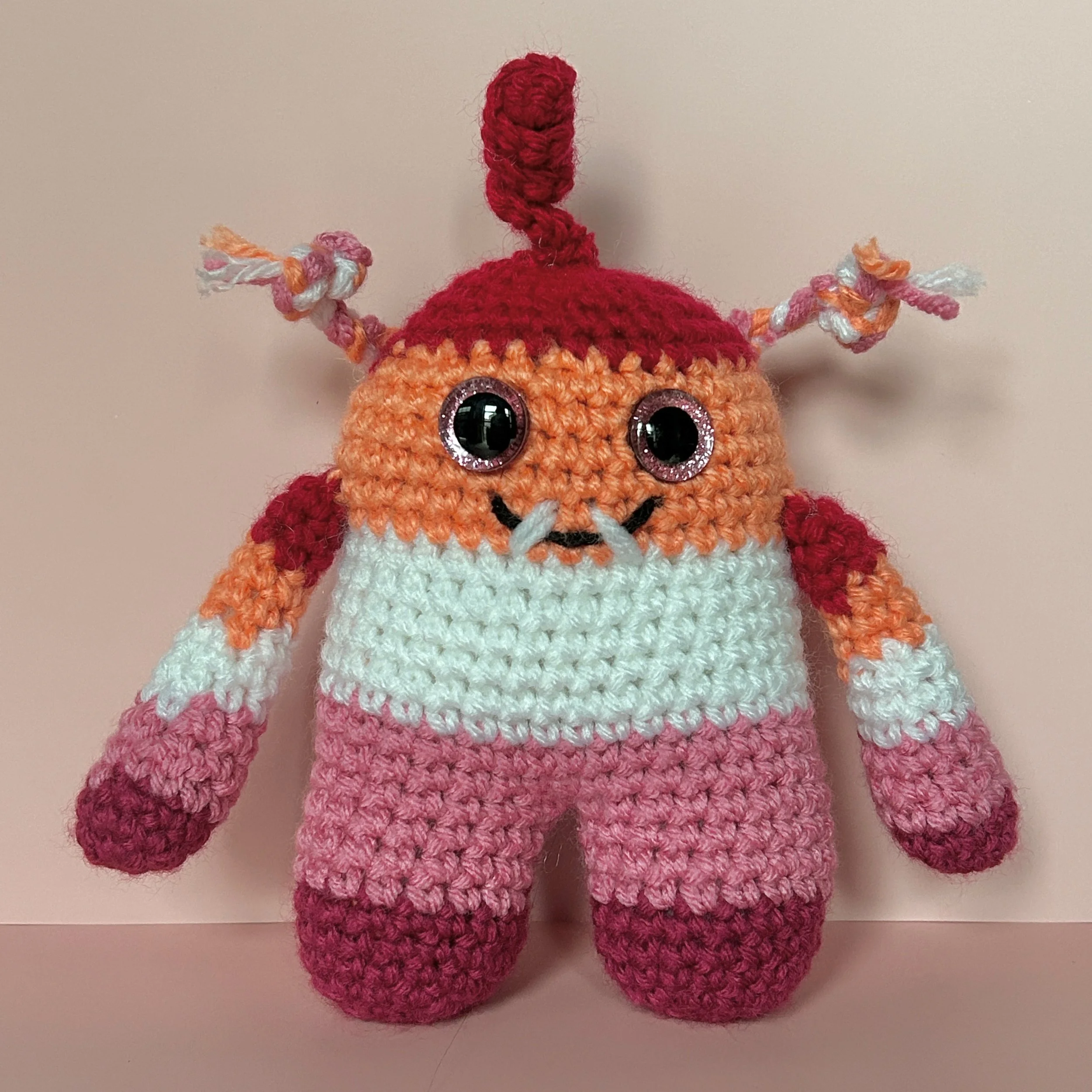 friendly dark orange, light orange, white, light pink, dark pink striped monster with braids and a squiggle hair pink googly eyes smiling with two teeth