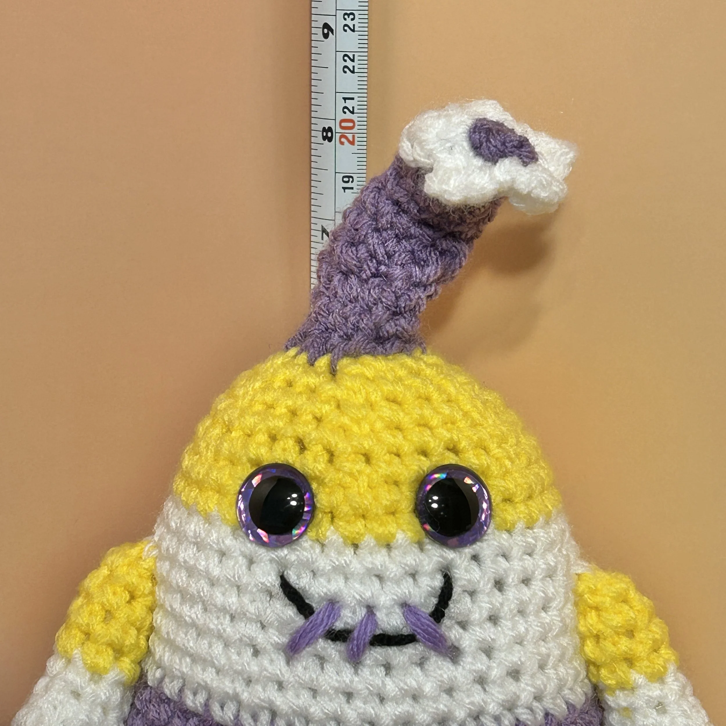Kye yellow white purple black striped monster with purple horn topped with white flower, purple googly eyes, smiling with 3 teeth in front of ruler body about 6 inches tall