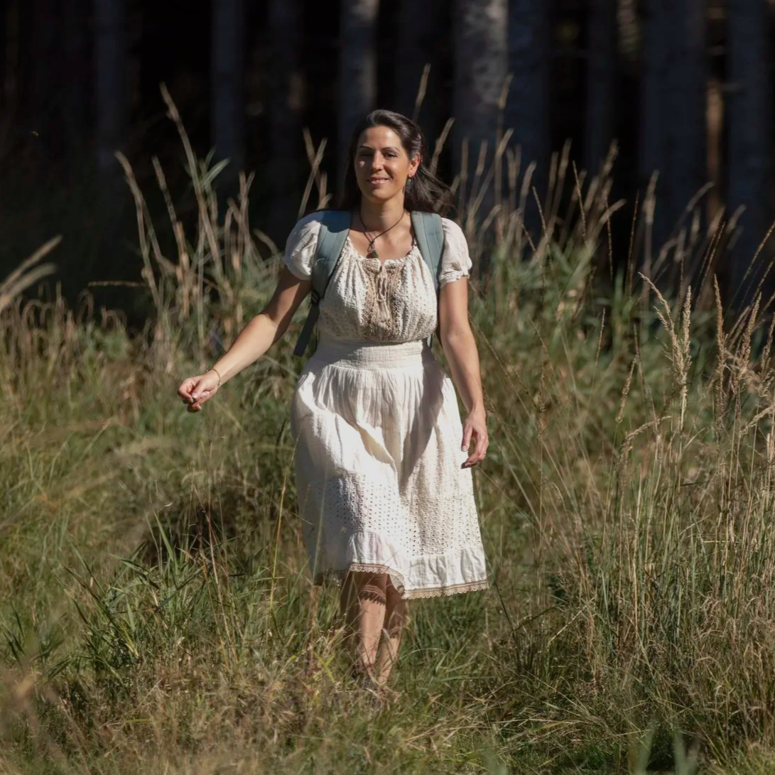 Eine junge Frau wandert durch eine Wiese im Wald bei Sonnenlicht, trägt ein weißes Kleid und einen Rucksack.