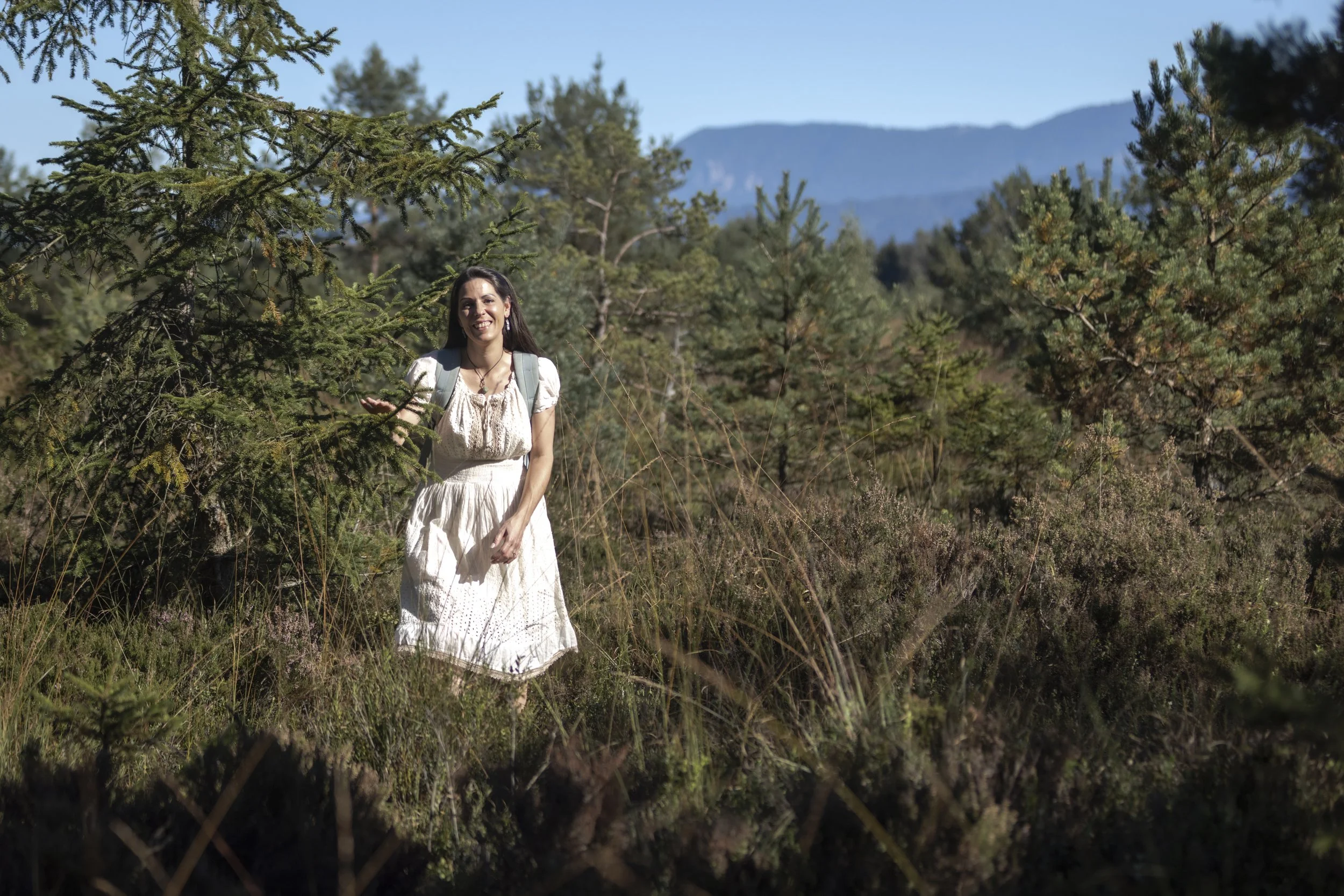 Junge Frau wandert durch einen Wald in den Bergen an einem sonnigen Tag, trägt ein weißes Kleid und einen Rucksack.