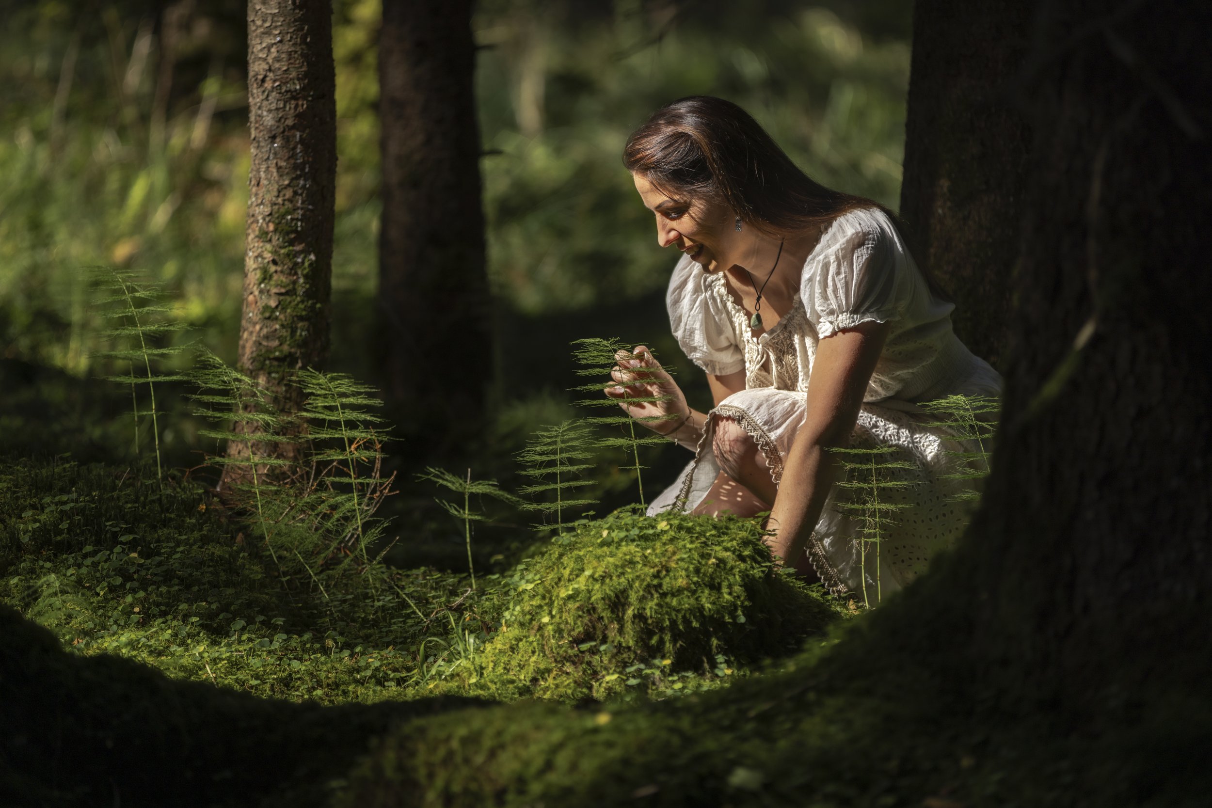 Eine lachende Frau in einem weißen Kleid, kniend im Wald, umgeben von grünen Pflanzen, die die Natur erkundet.