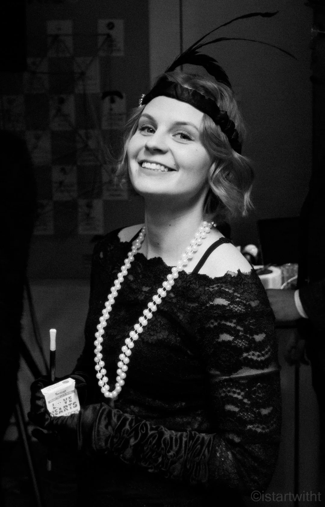 A woman smiling at a costume party, dressed in 1920s vintage attire with a feather headpiece, pearl necklace, lace dress, and gloves, holding a fake cigarette candy box.
