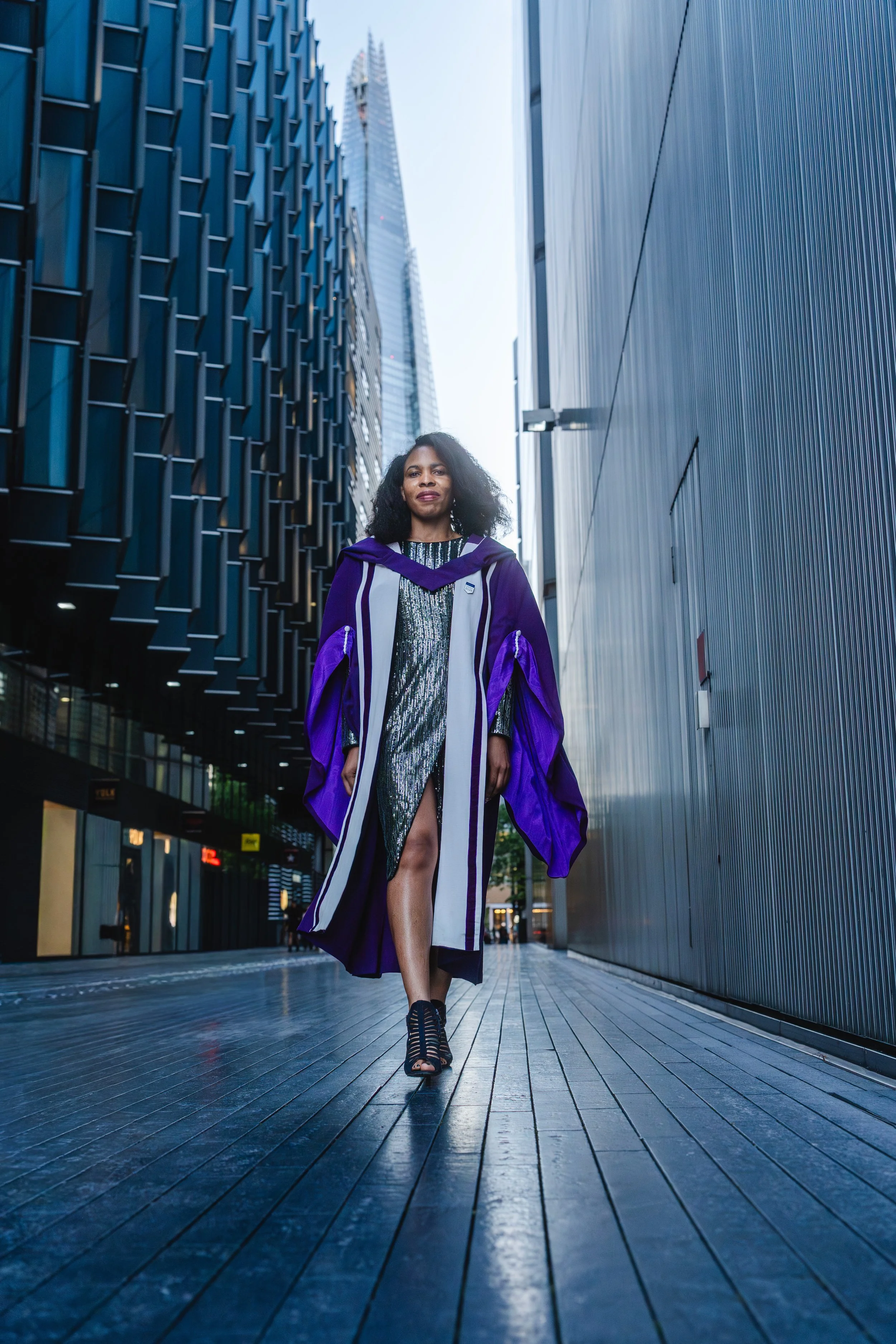 A black woman in a shiny, metallic dress and a purple and white graduation gown walking down a London street with the Shard in the background.