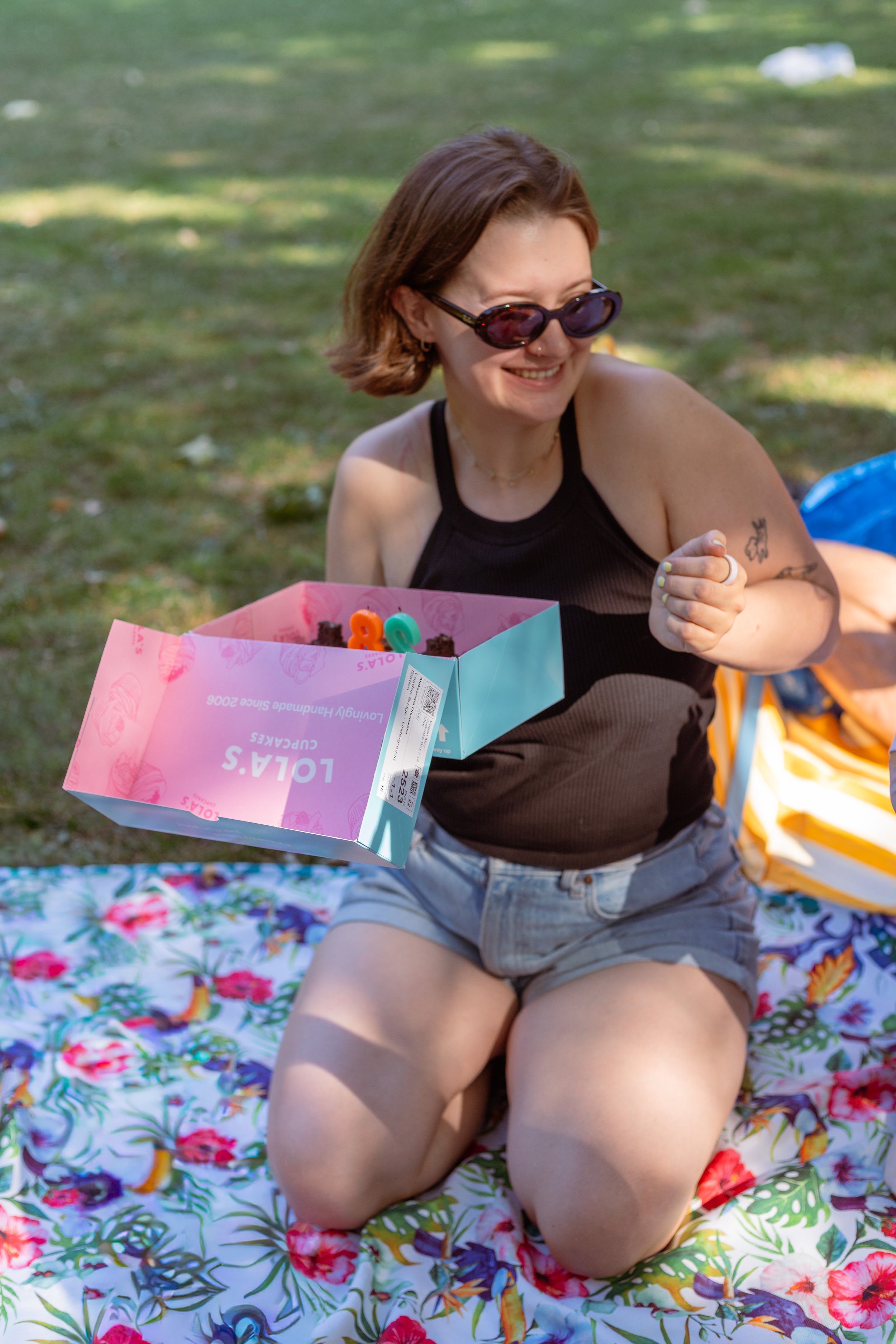 A white woman laughing and kneeling outdoors on a picnic blanket. She wears oval sunglasses and is holding her birthday cupcakes. A candid portrait.