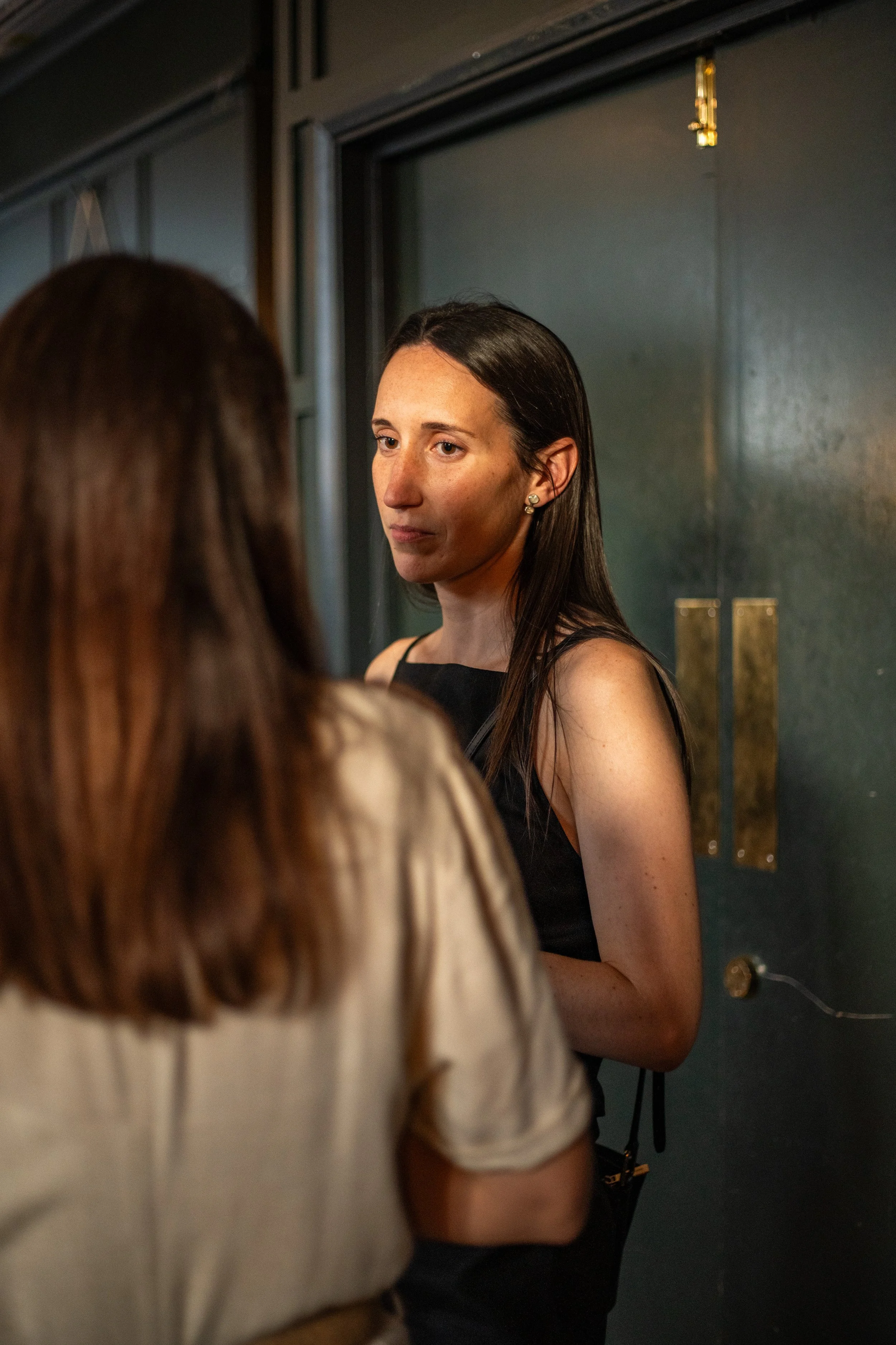 A woman with long dark hair and earrings stands against a dark sage green wall with two brass plaques Her face is lit up golden by the evening sun and she is engaged in a conversation with another person in the foreground.