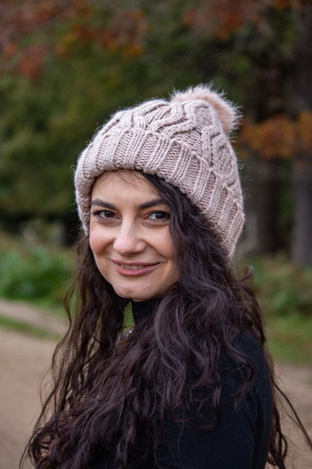 A woman with long curly dark hair smiling outdoors, wearing a beige knitted beanie with a pom-pom in fall or winter.