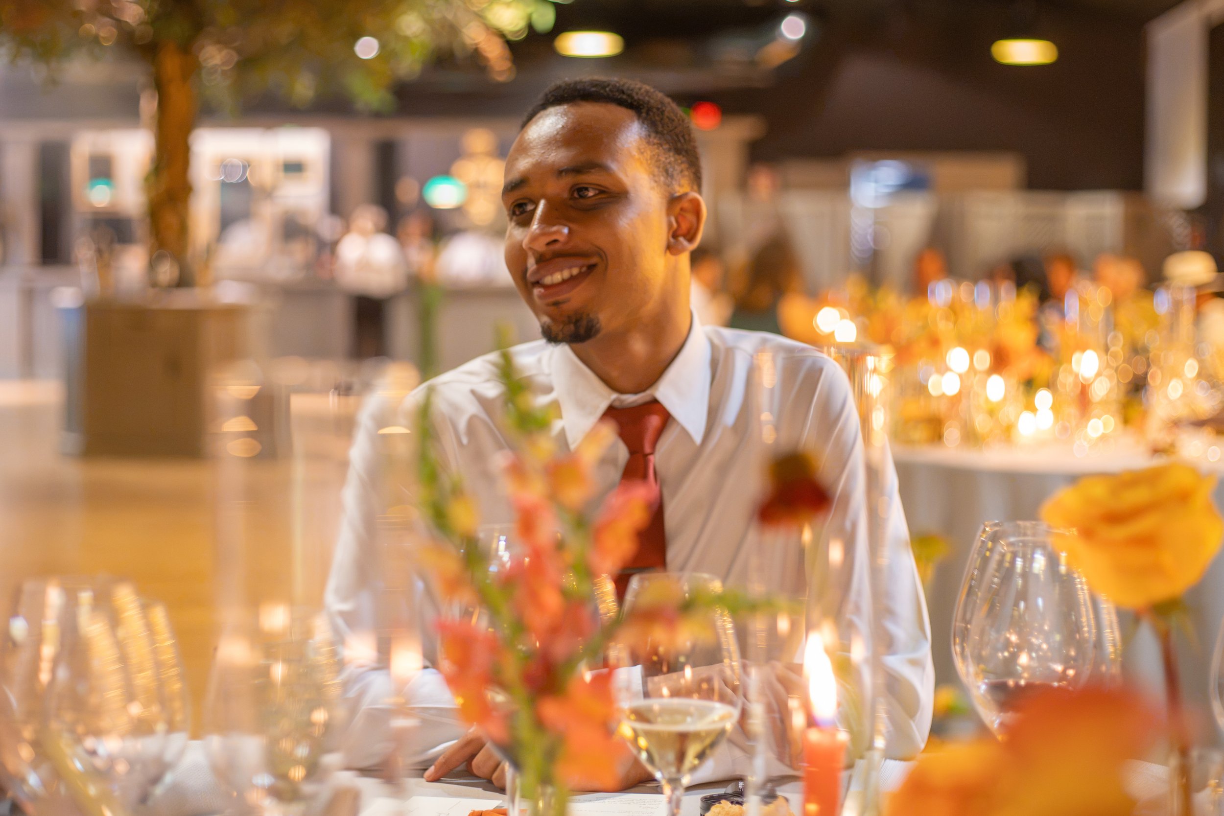 A smiling young black man in a white dress shirt and red tie sitting at a table decorated for a wedding, with flowers and glassware, in a warmly lit conservatory hall.