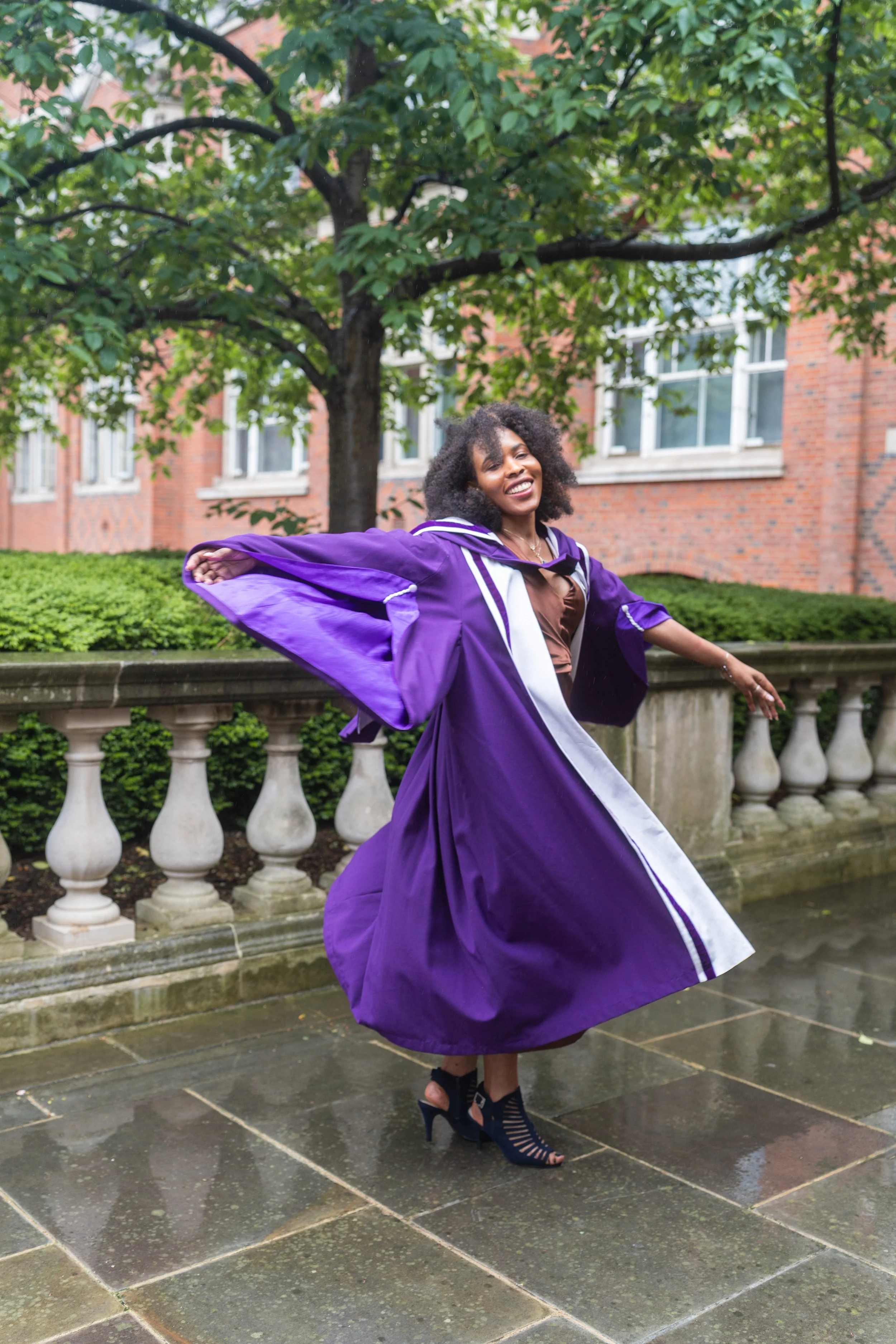 A black woman in a purple and white graduation gown and heels is twirling outdoors on a wet slate pavement, with a brick building and green trees in the background.