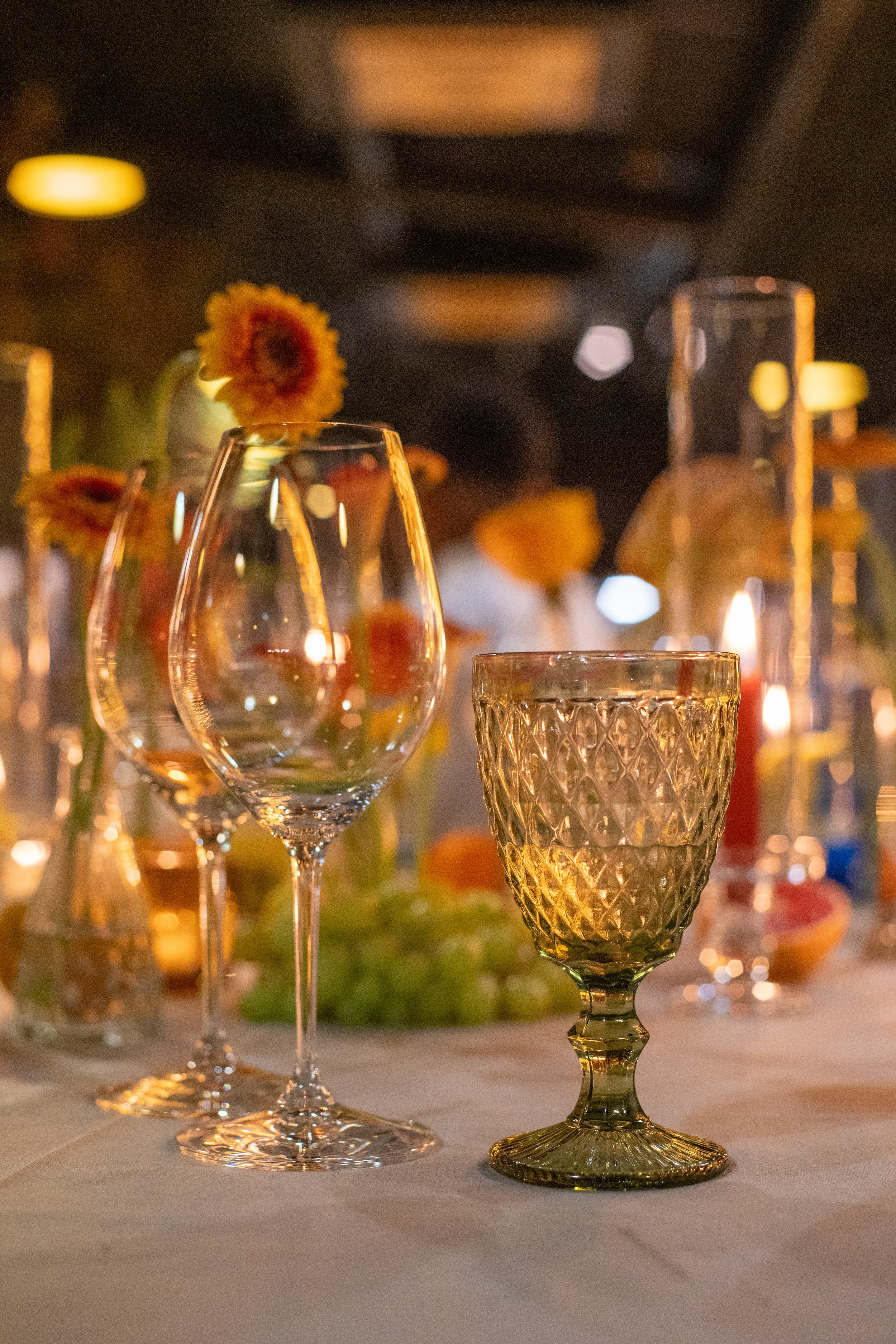 A close-up of various empty glasses on a table, with some decorated with flowers, set in a warmly lit environment, possibly for a celebration or event.