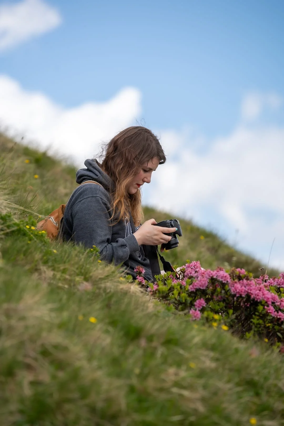 A white female photographer sitting on a grassy hillside, taking a picture of flowers with a digital camera, under a partly cloudy sky.