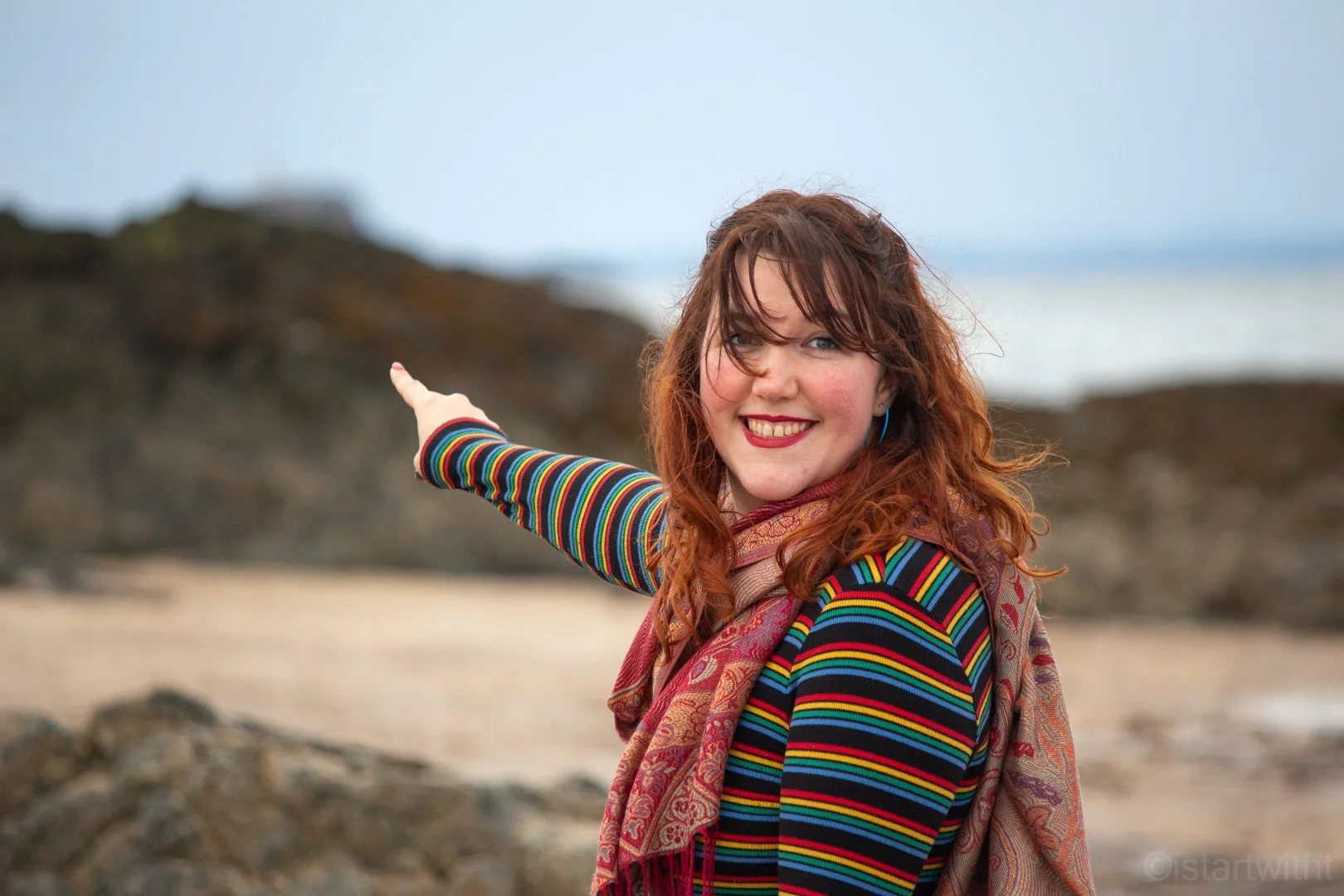 A white woman with red hair wearing a colorful striped long-sleeve shirt and a patterned scarf, smiling, and pointing towards the distant landscape on a cloudy day.