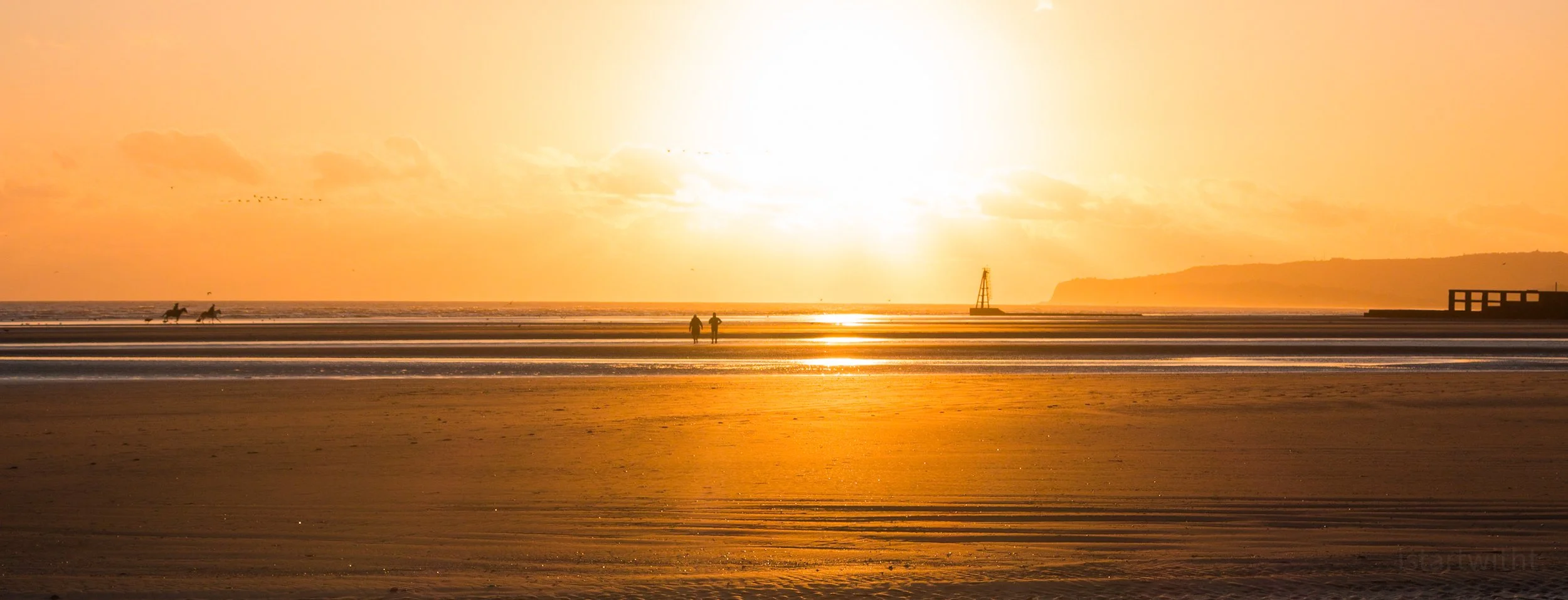 Sunset over the Camber Sands beach with two people walking, horses in the distance, and a lighthouse on the horizon.